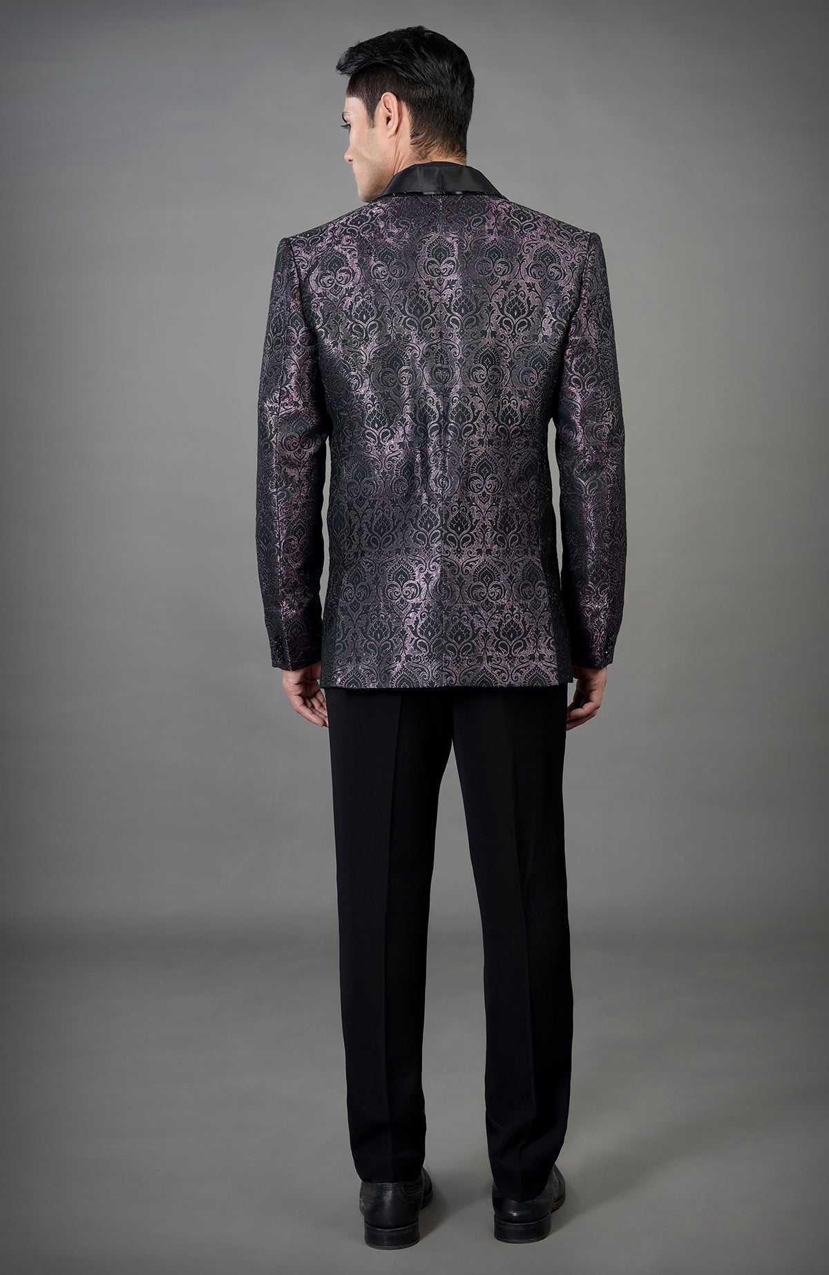A man from behind wears a black woven brocade tuxedo with black pants.