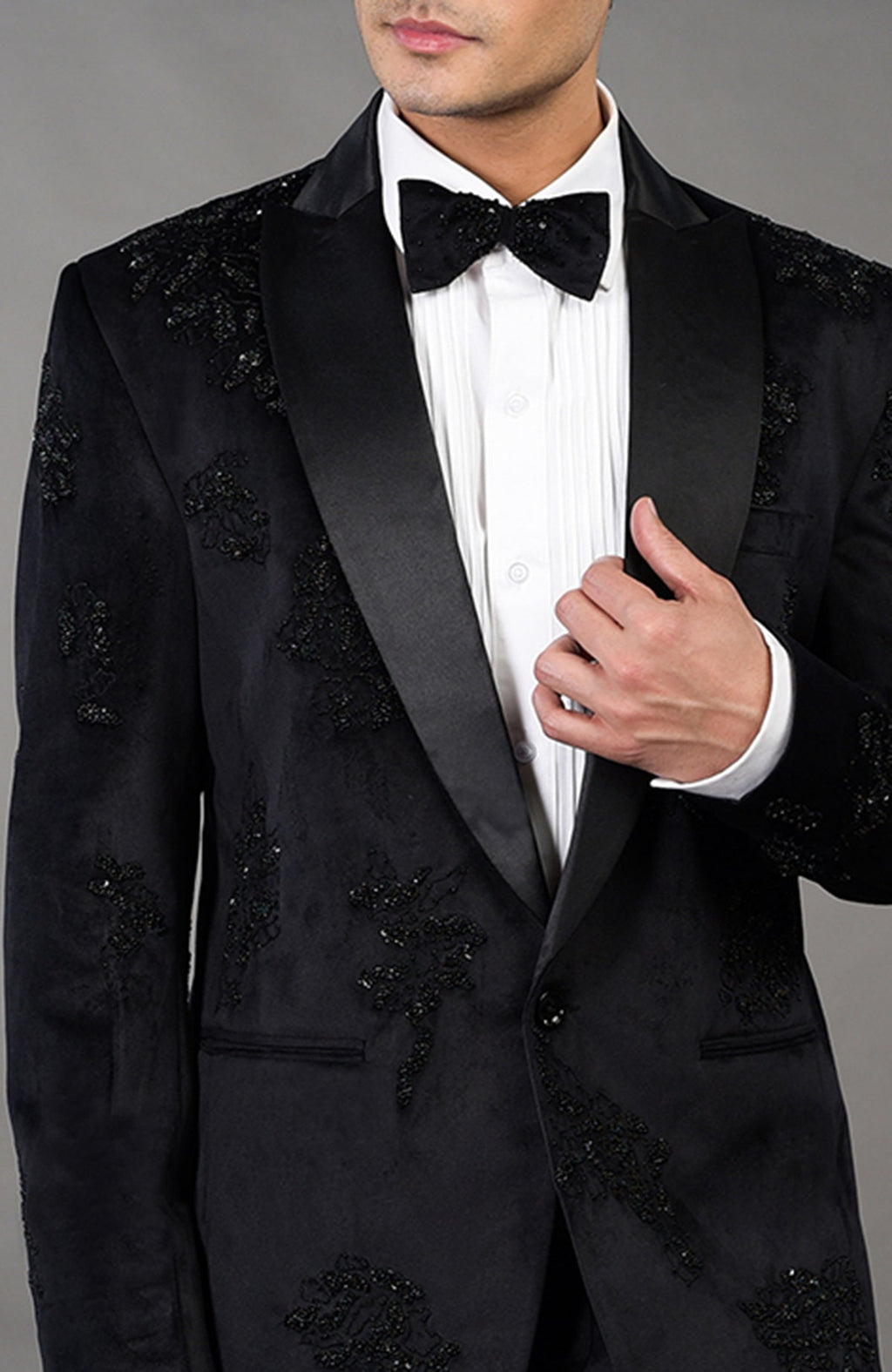 A man models a black tuxedo with intricate beadwork and a white pleated shirt.