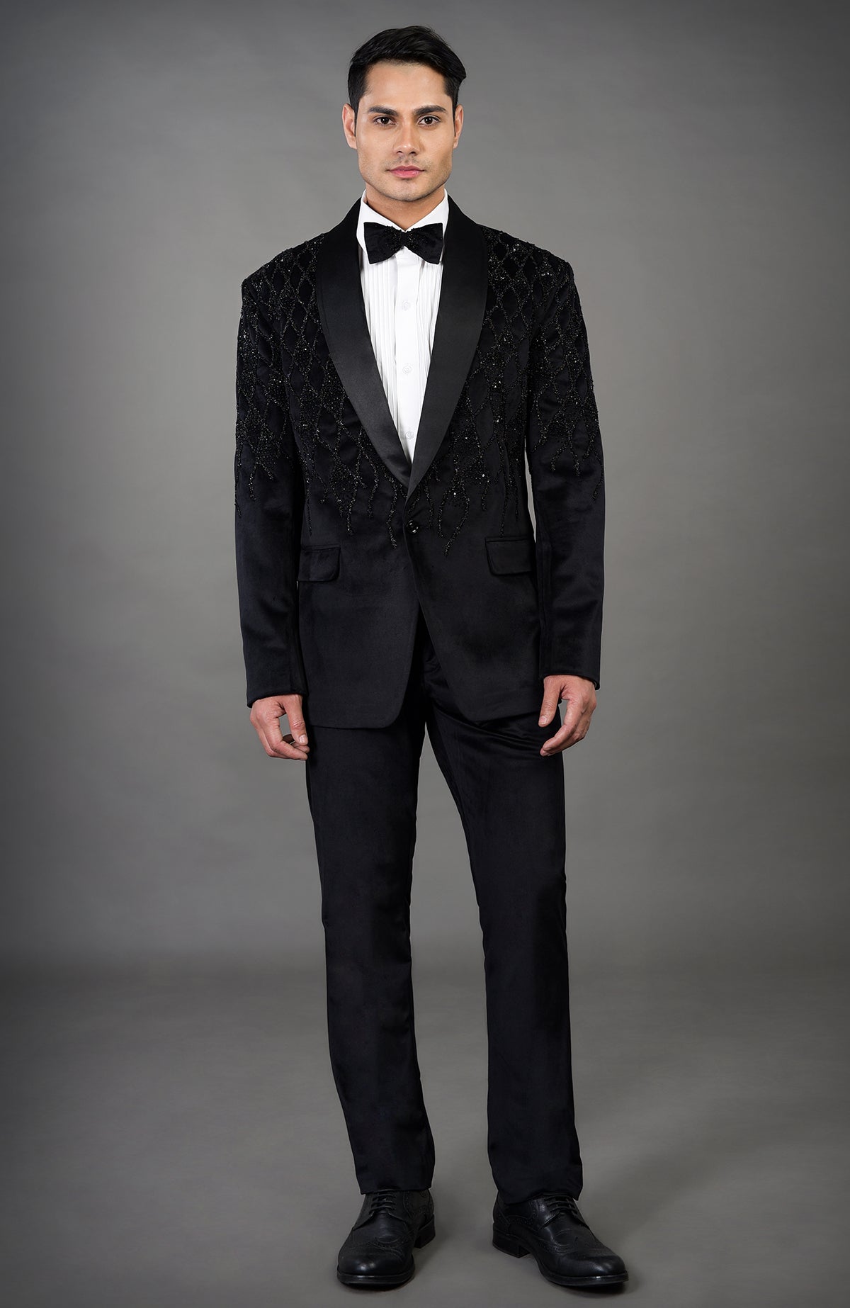 A man models a black velvet tuxedo with beaded embroidery and a white shirt.