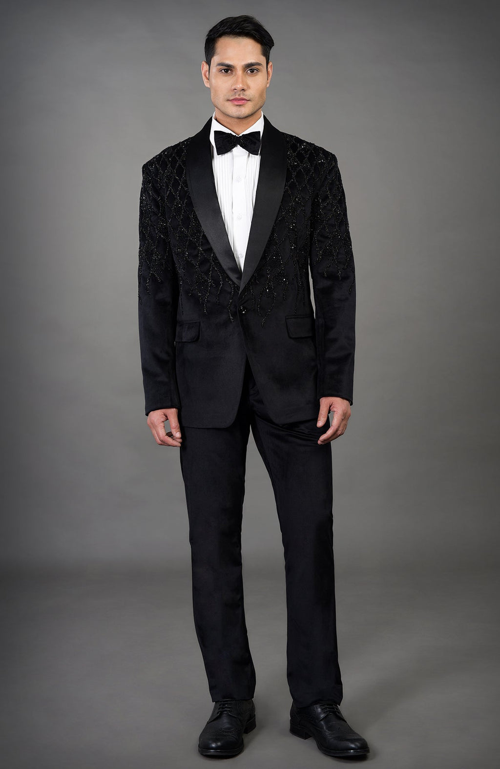 A man models a black velvet tuxedo with beaded embroidery and a white shirt.