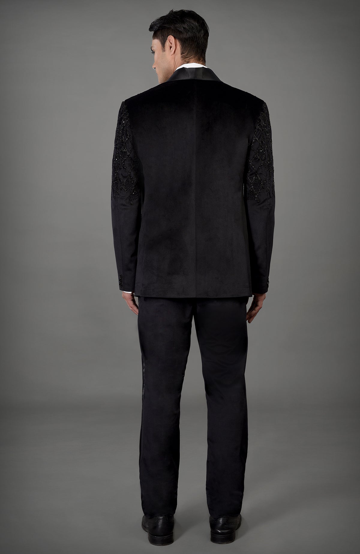 A man from behind wears a black velvet tuxedo with embroidered sleeves.
