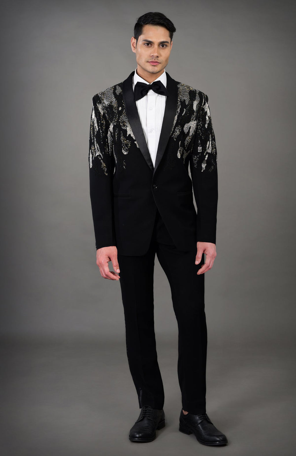 A man models a black tuxedo with a sequined abstract pattern on the shoulders and sleeves.