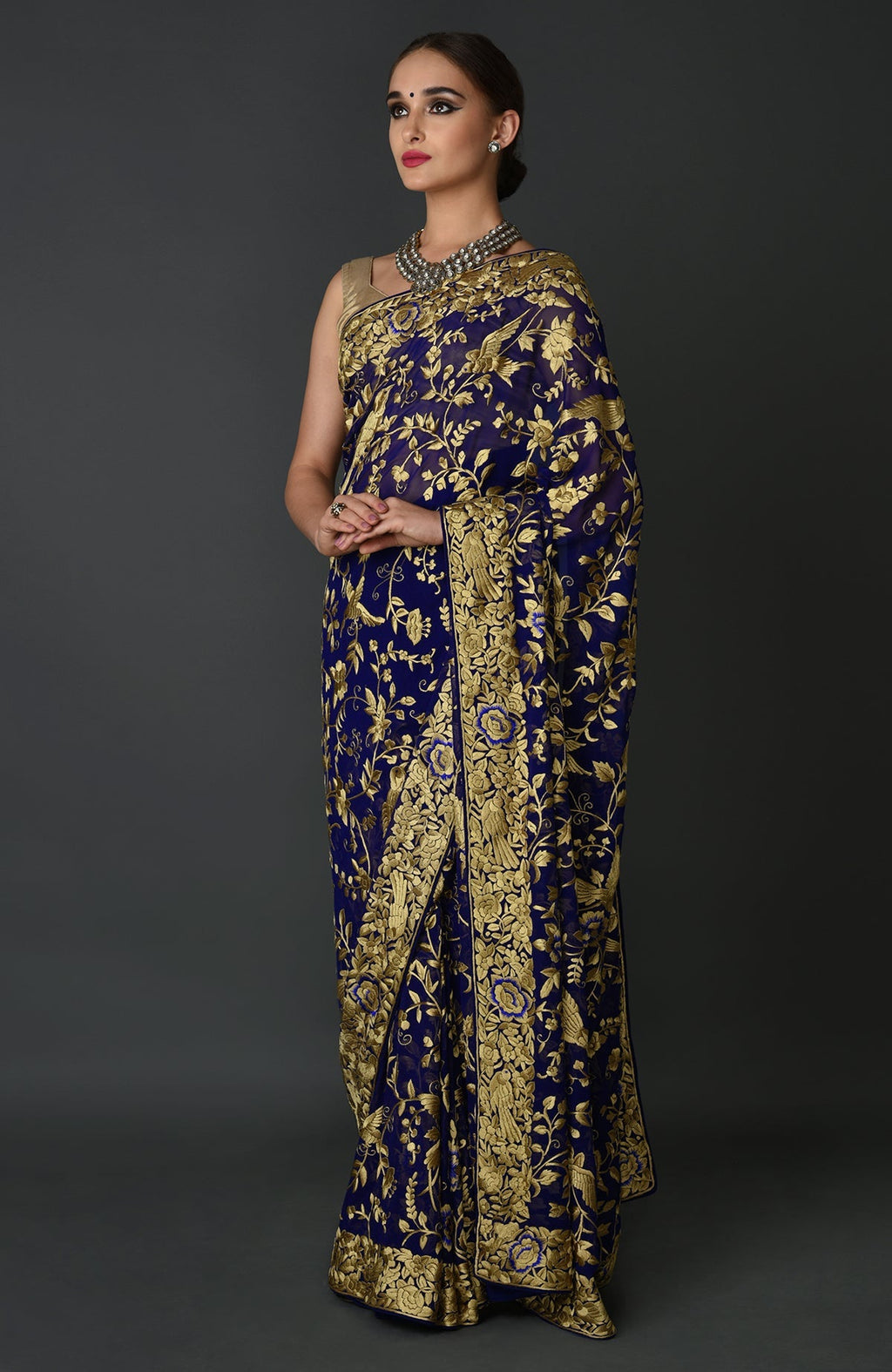 A woman models an indigo blue Parsi Gara hand-embroidered saree with gold floral patterns.