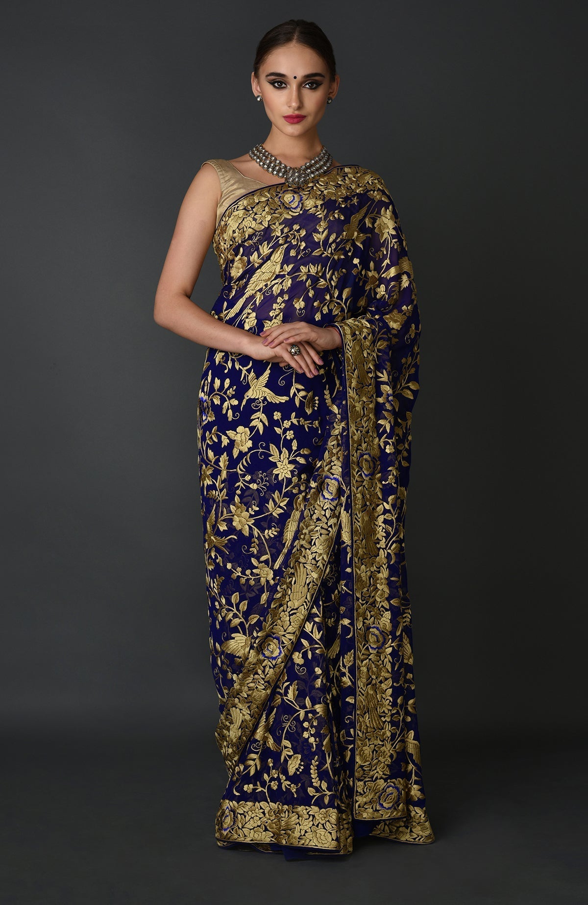 A woman models an indigo blue Parsi Gara hand-embroidered saree with gold floral patterns.
