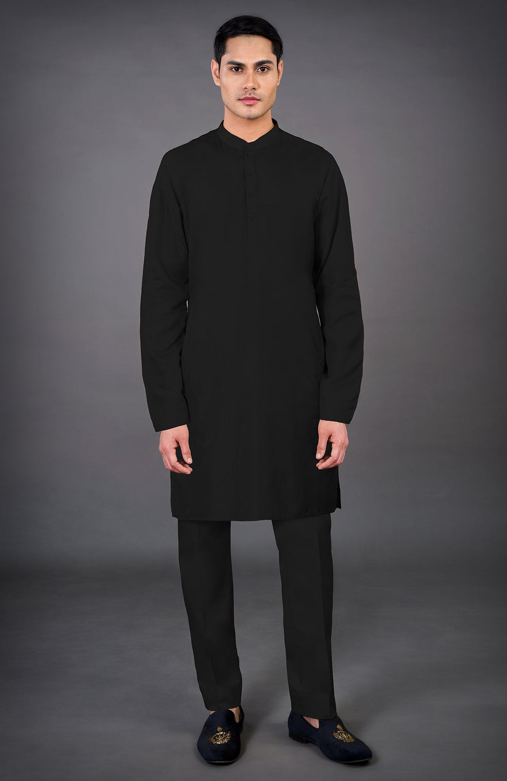 A man stands wearing a black kurta and matching pants with embroidered loafers.