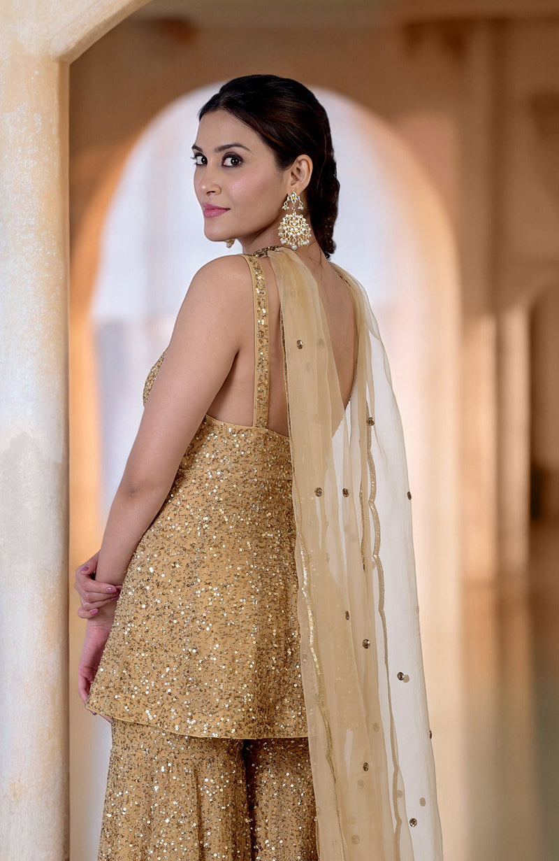 A woman in a gold sequin sharara set with a sheer dupatta looks over her shoulder.