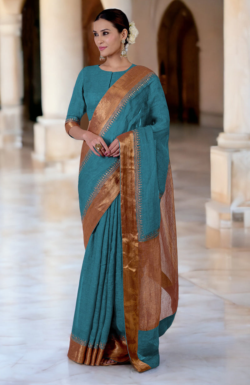 A woman models a teal linen saree with gold embroidered borders and a matching blouse.