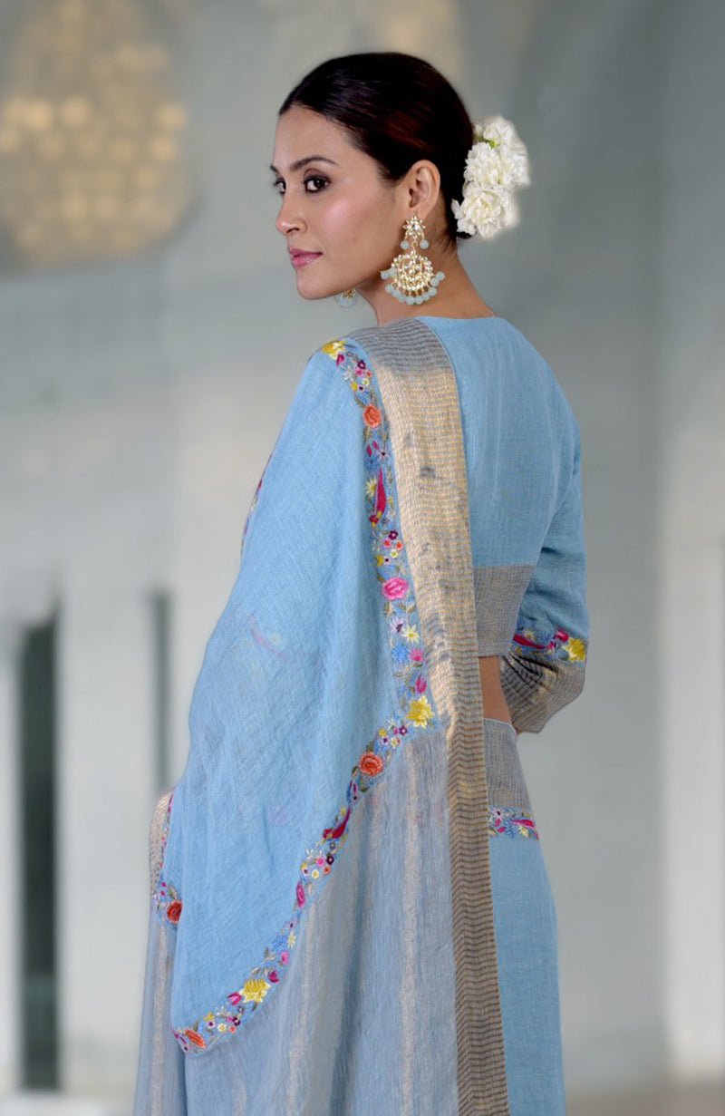 A woman models a cerulean blue linen saree with Parsi Gara embroidery and zari details.
