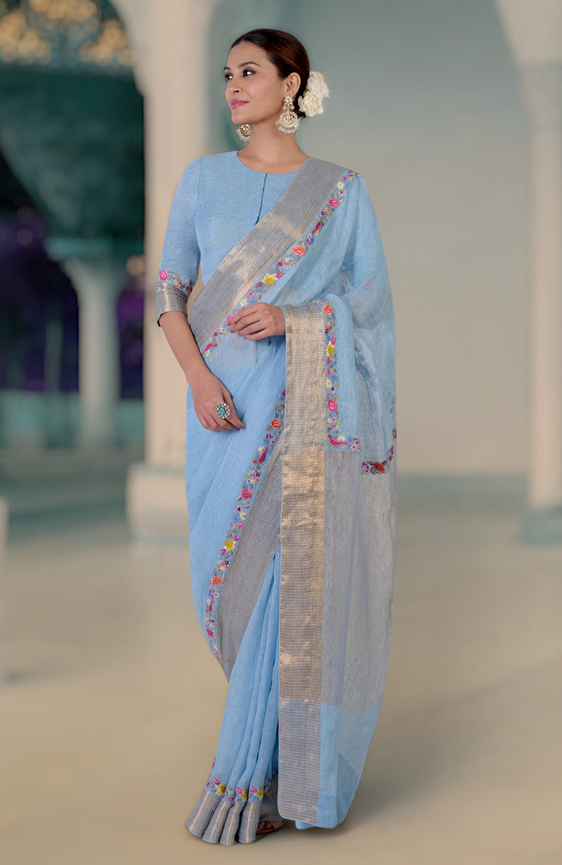 A woman models a cerulean blue linen saree with Parsi Gara embroidery and zari details.
