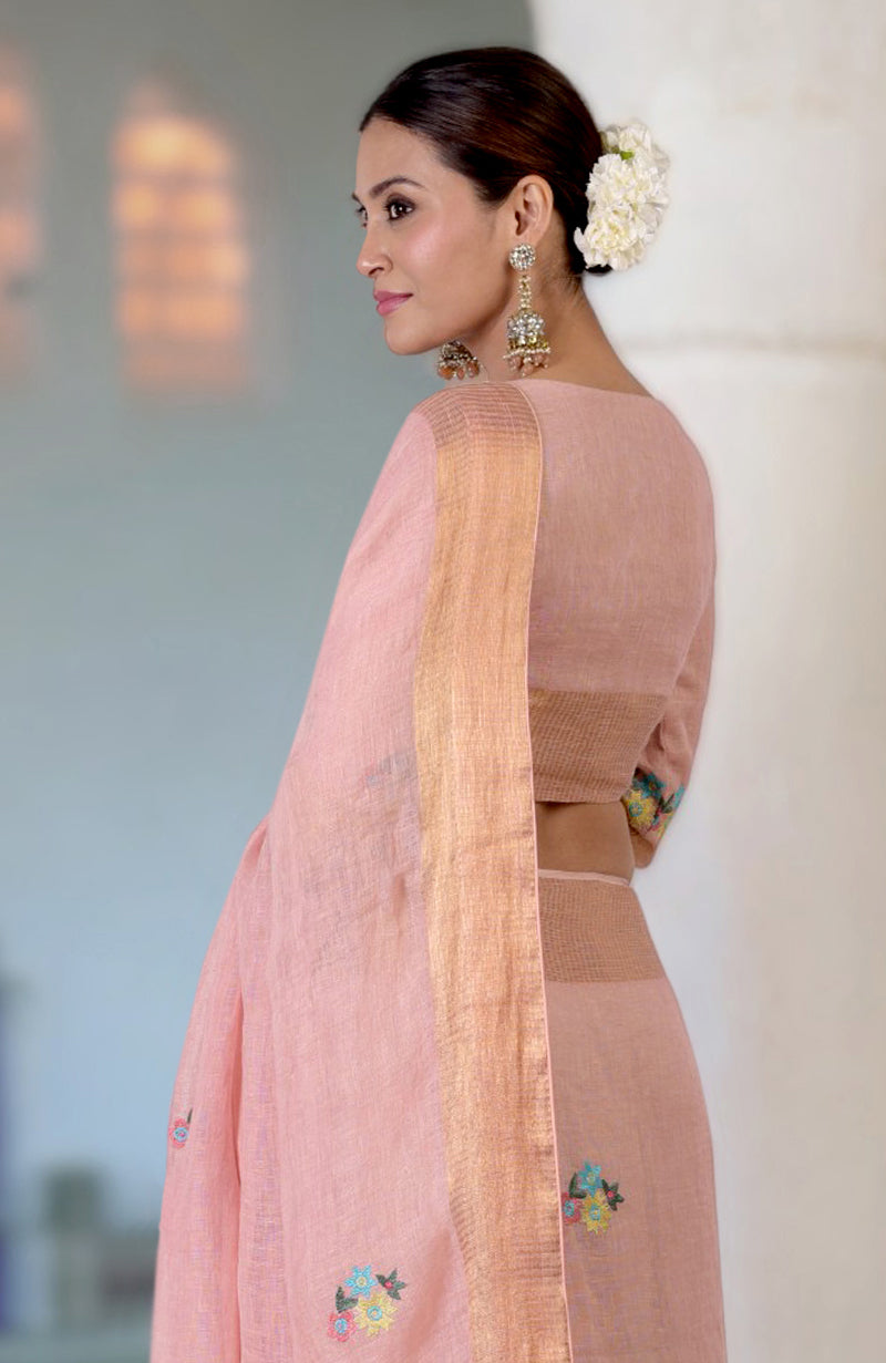 A woman in a pink linen saree with floral embroidery and a gold zari border.