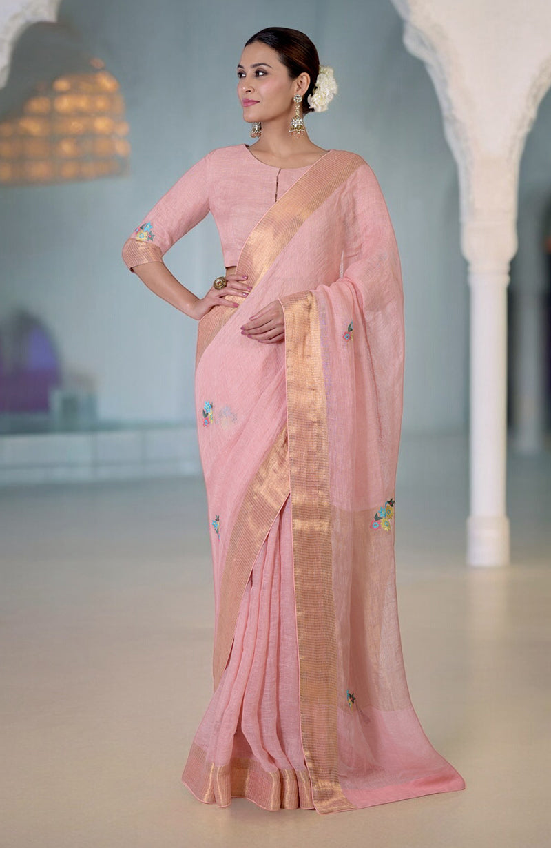 A woman models a pink linen saree with gold zari borders and colorful floral embroidery.