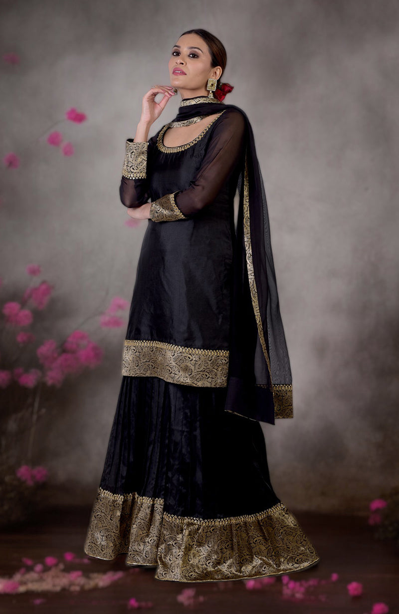 A woman models a black and gold Banarasi embroidered sharara set with a red flower in her hair.