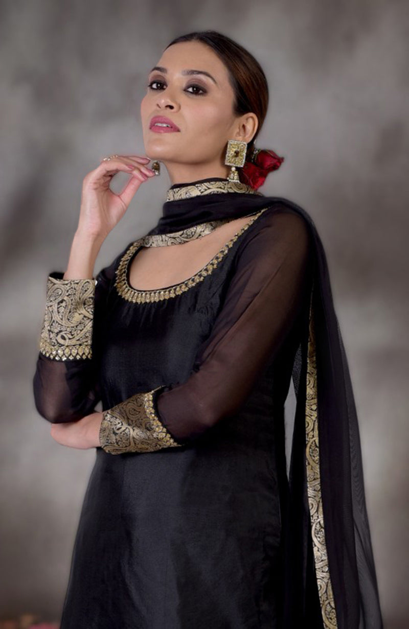 A woman models a black and gold Banarasi embroidered sharara set with a red rose in her hair.