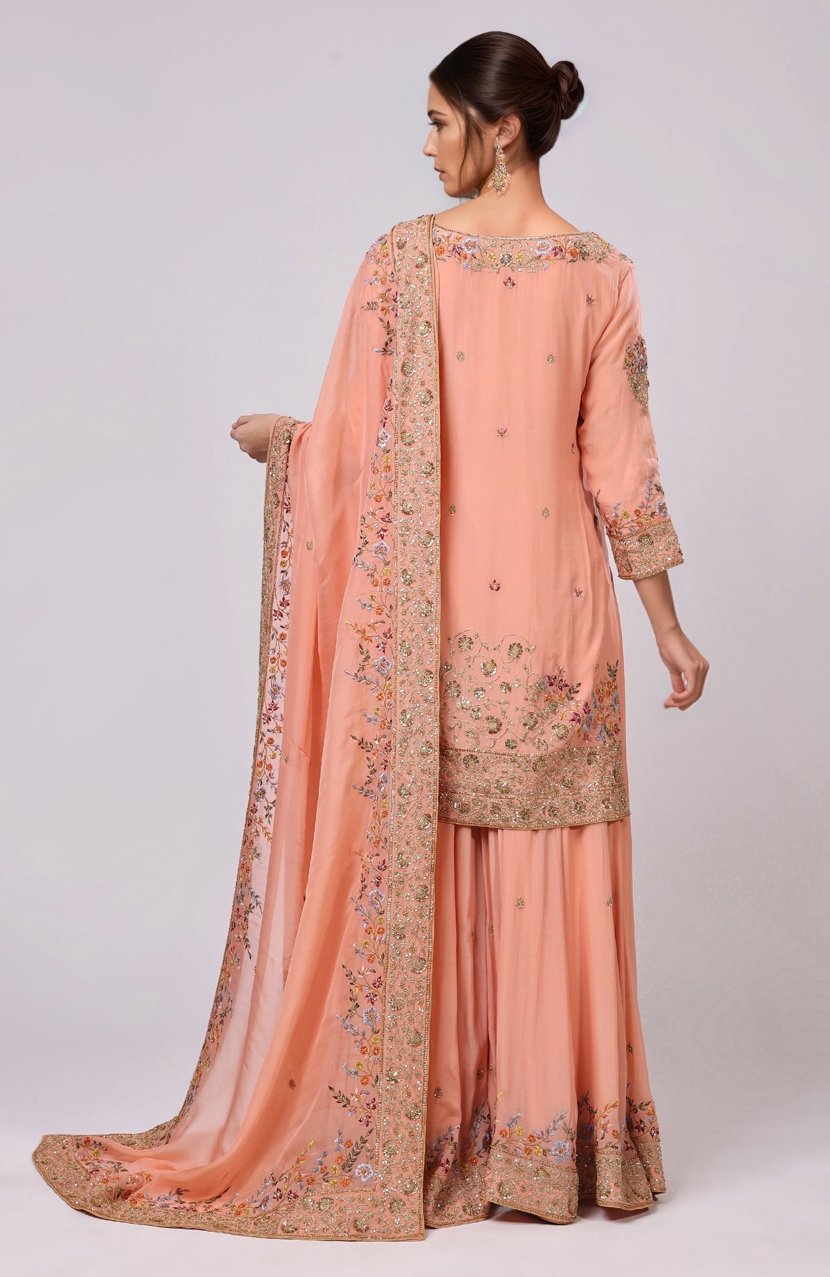 A woman models a salmon sharara set with intricate embroidery and a matching dupatta.