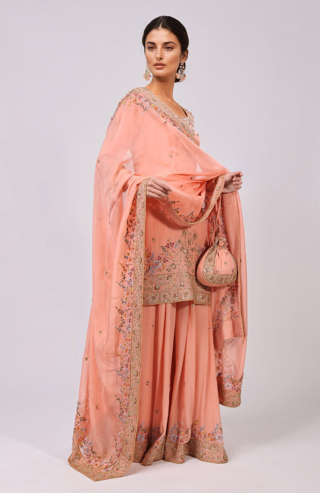 A model wears a salmon sharara set with intricate embroidery and a matching potli bag.