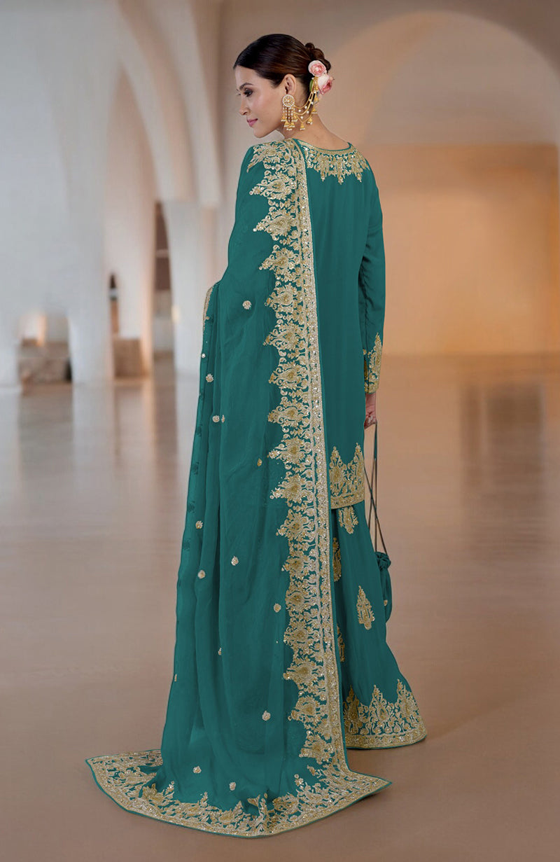 A woman in a teal sharara set with gold embroidery stands facing away.