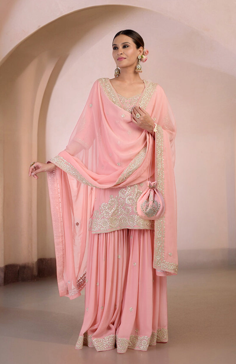 A woman in a pink sharara set with gold embroidery and a matching potli bag.