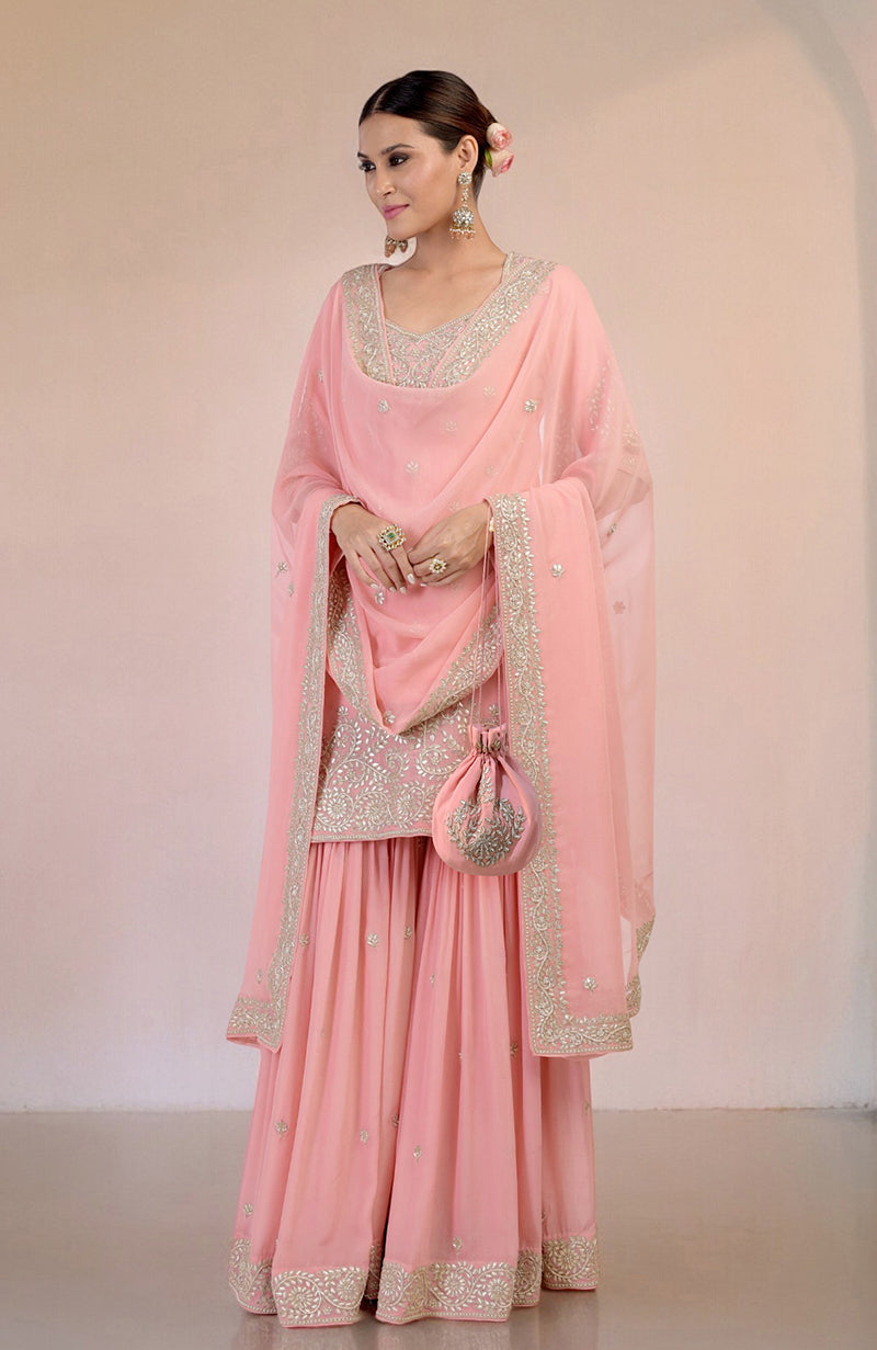 A woman in a pink sharara set with intricate embroidery and a matching potli bag.
