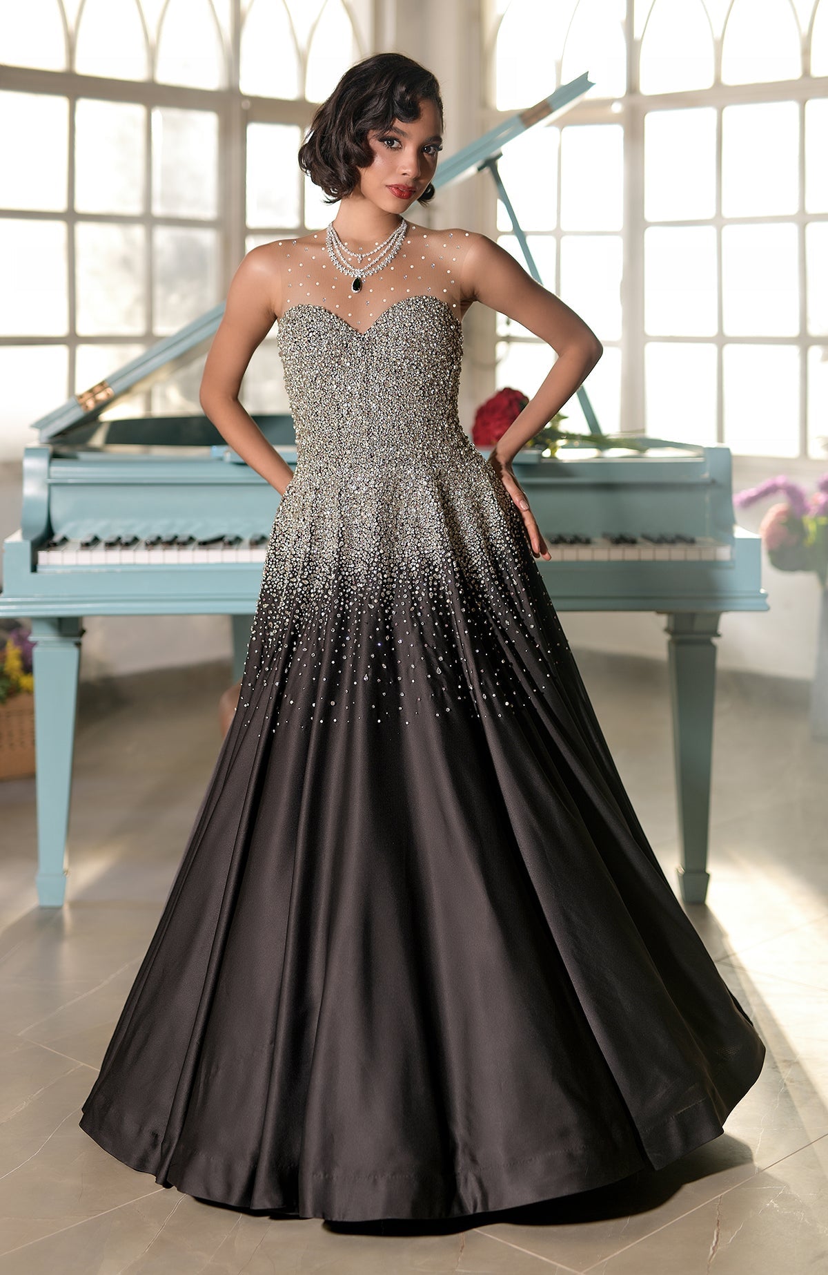 A woman in a black and silver crystal-embroidered princess gown stands by a light blue piano.