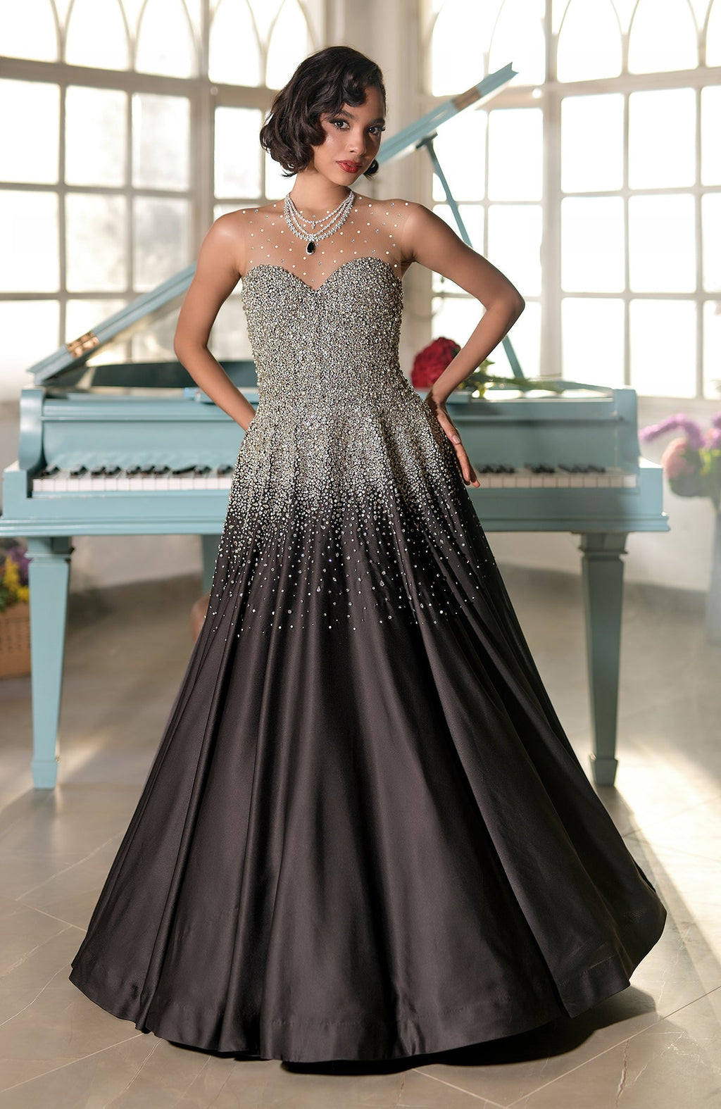 A woman in a black and silver crystal-embroidered princess gown stands by a light blue piano.