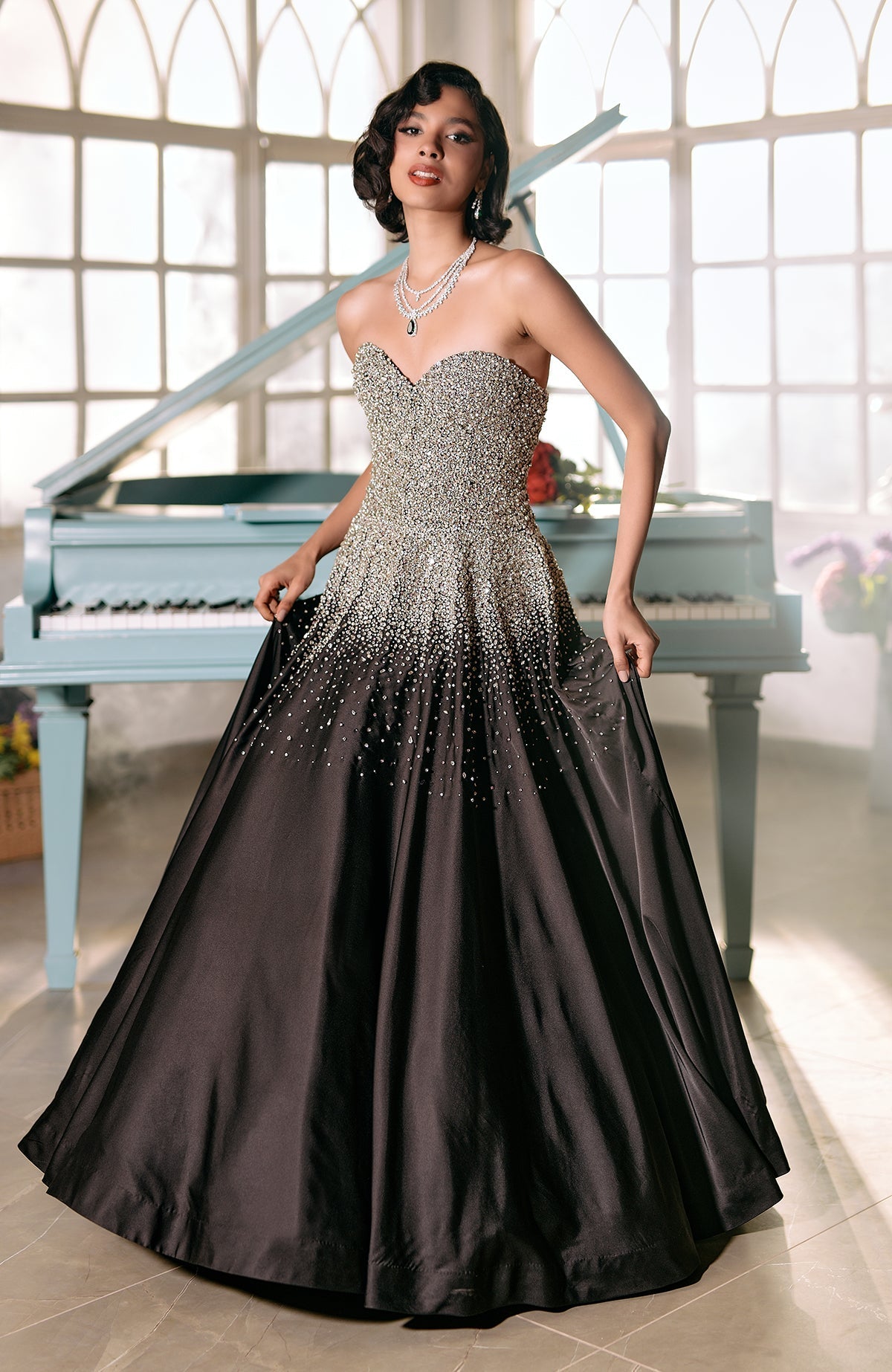 A woman in a black and crystal-beaded princess gown stands by a light blue piano.