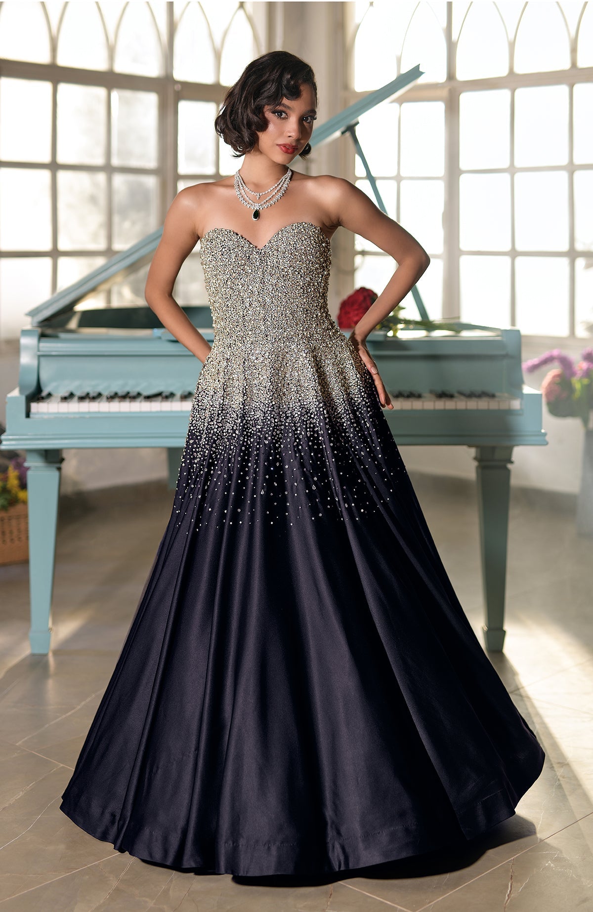 A woman in a black and gold crystal-embroidered strapless gown stands by a piano.