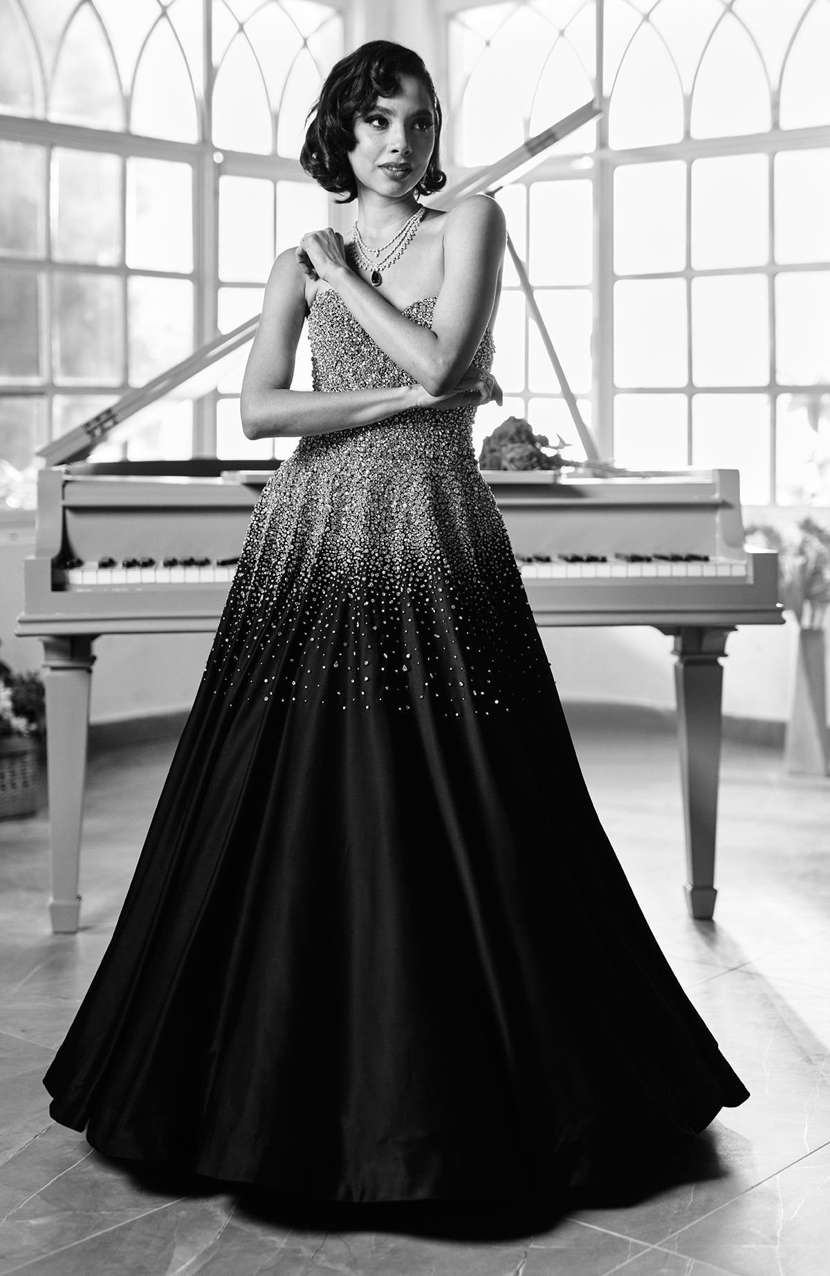 A woman in a black crystal hand-embroidered ball gown stands in front of a piano.