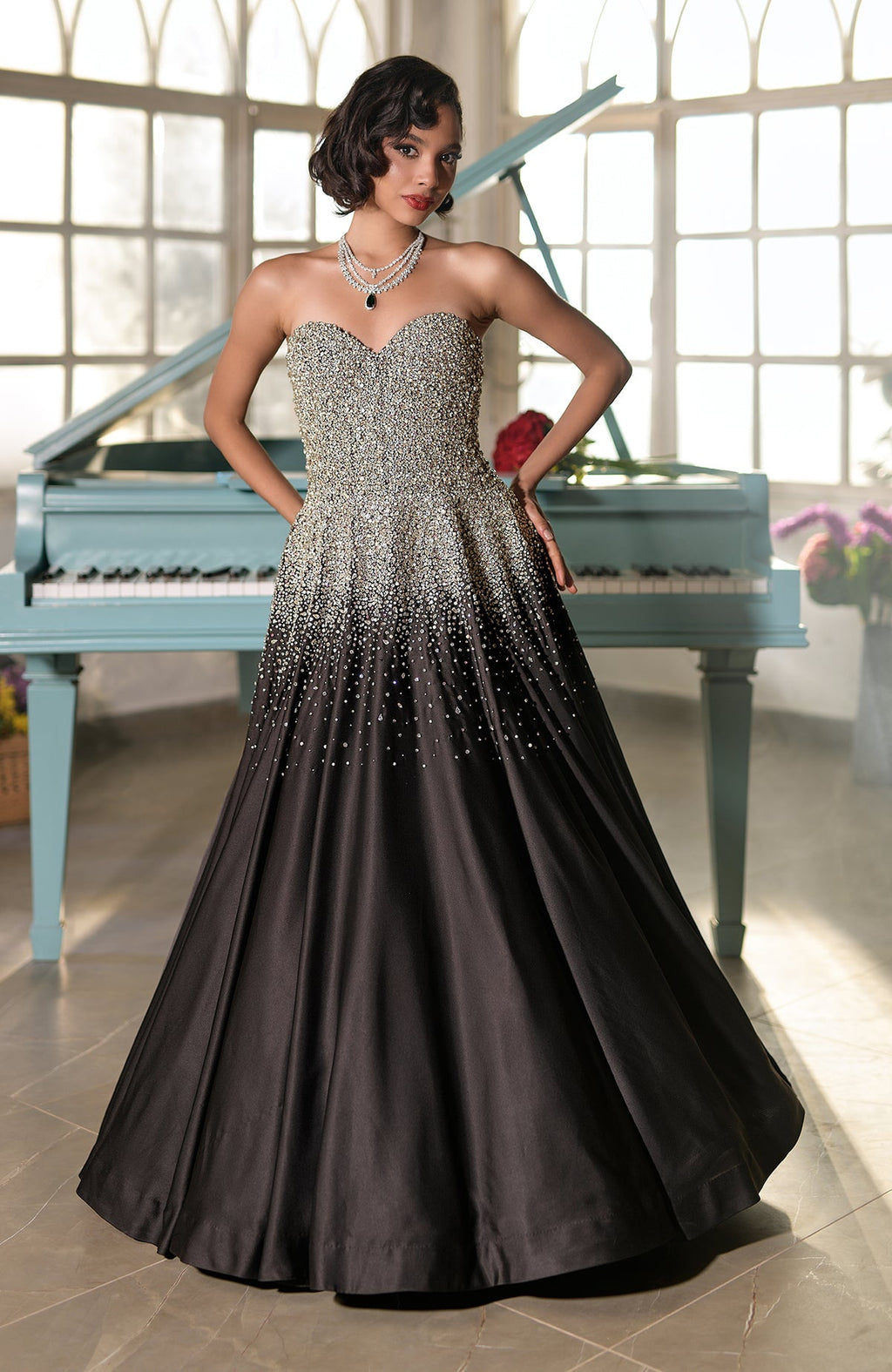 A woman in a black crystal hand-embroidered ball gown stands in front of a piano.