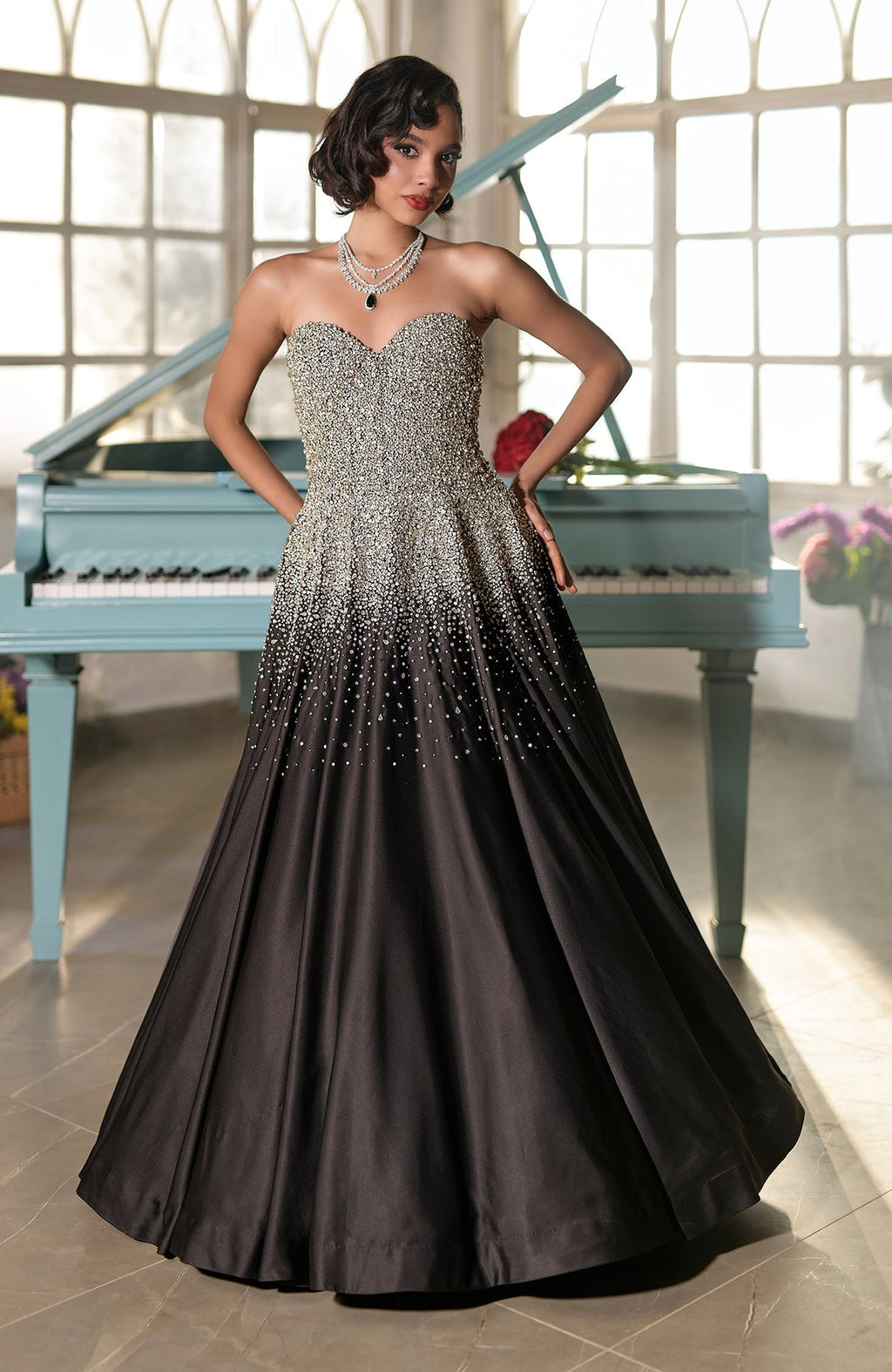 A woman in a black and gold crystal-embroidered princess gown stands by a piano.