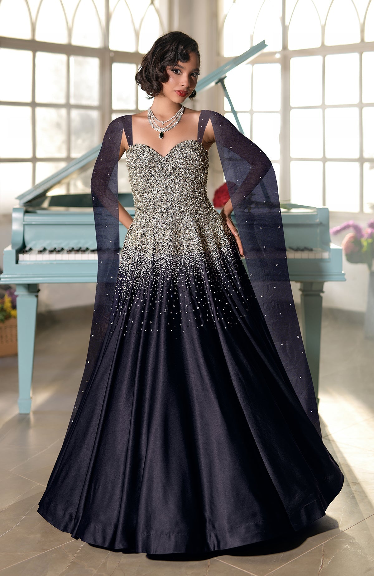 A woman in a black and gold embellished gown with sheer sleeves stands by a piano.