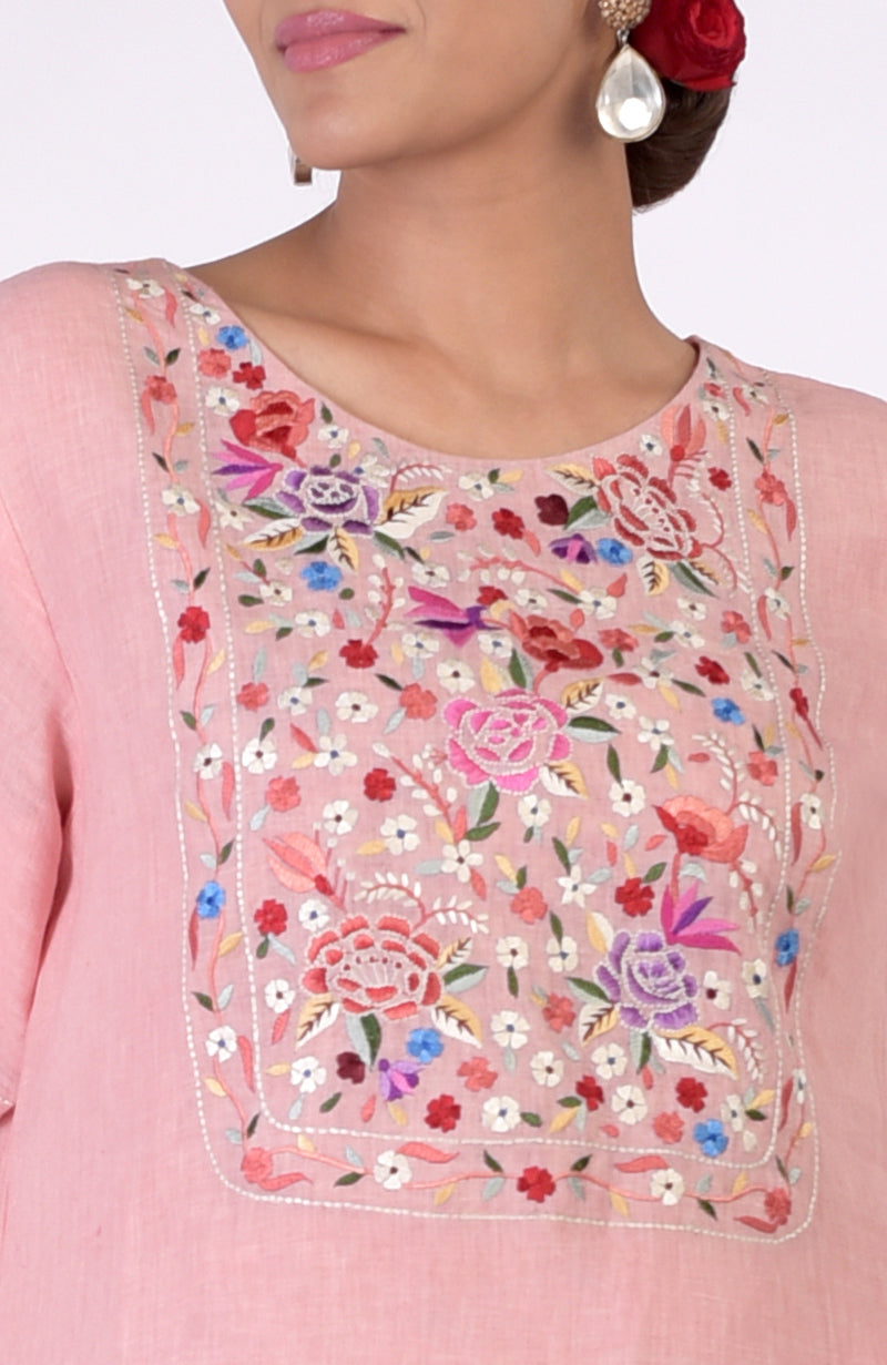 A woman wears a pink linen kurta with colorful floral Parsi Gara embroidery on the neckline.