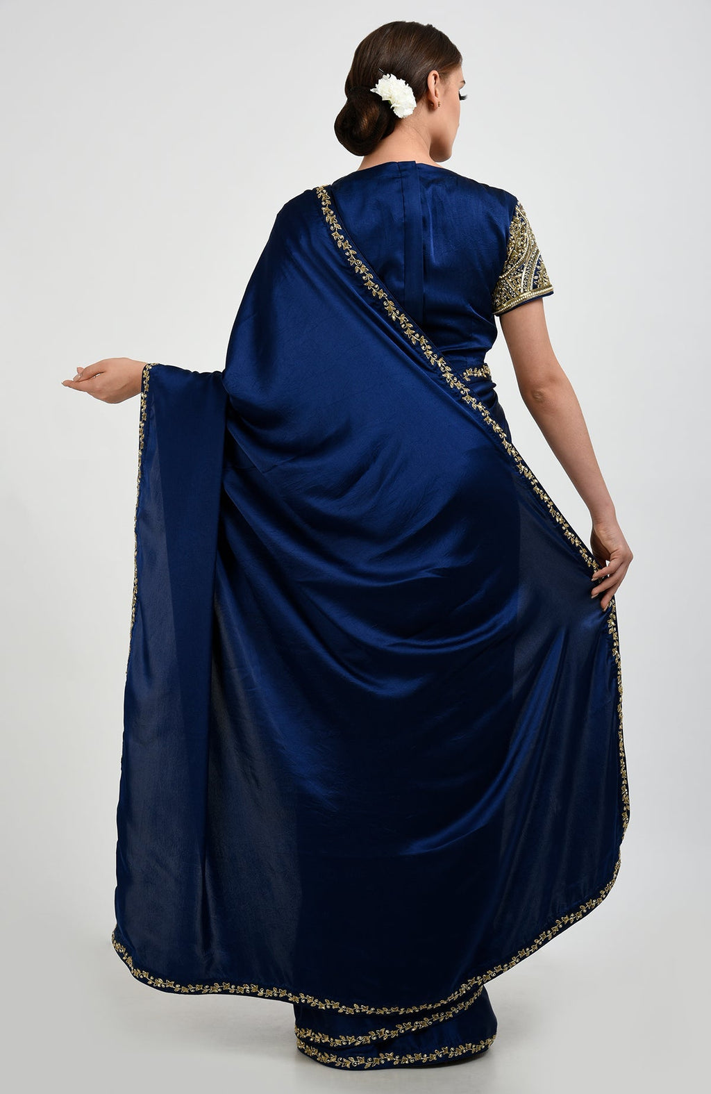 A woman models a sapphire blue saree with gold embroidery and a white flower in her hair.
