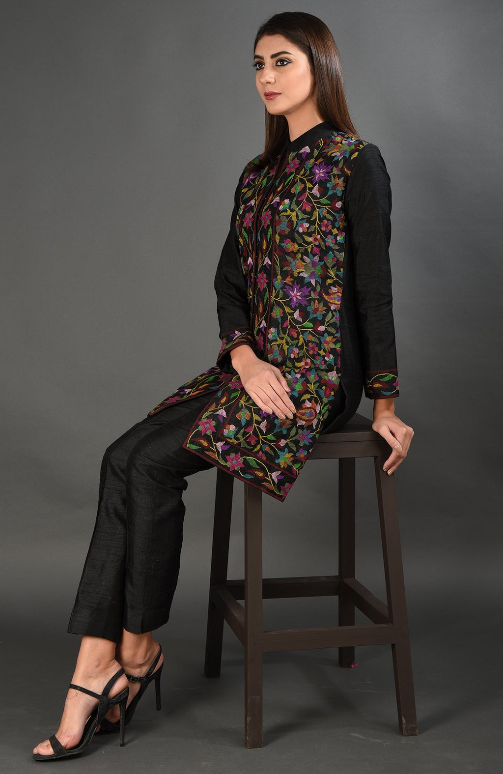 A woman sits wearing a black Kashmir Kani art embroidered jacket set.