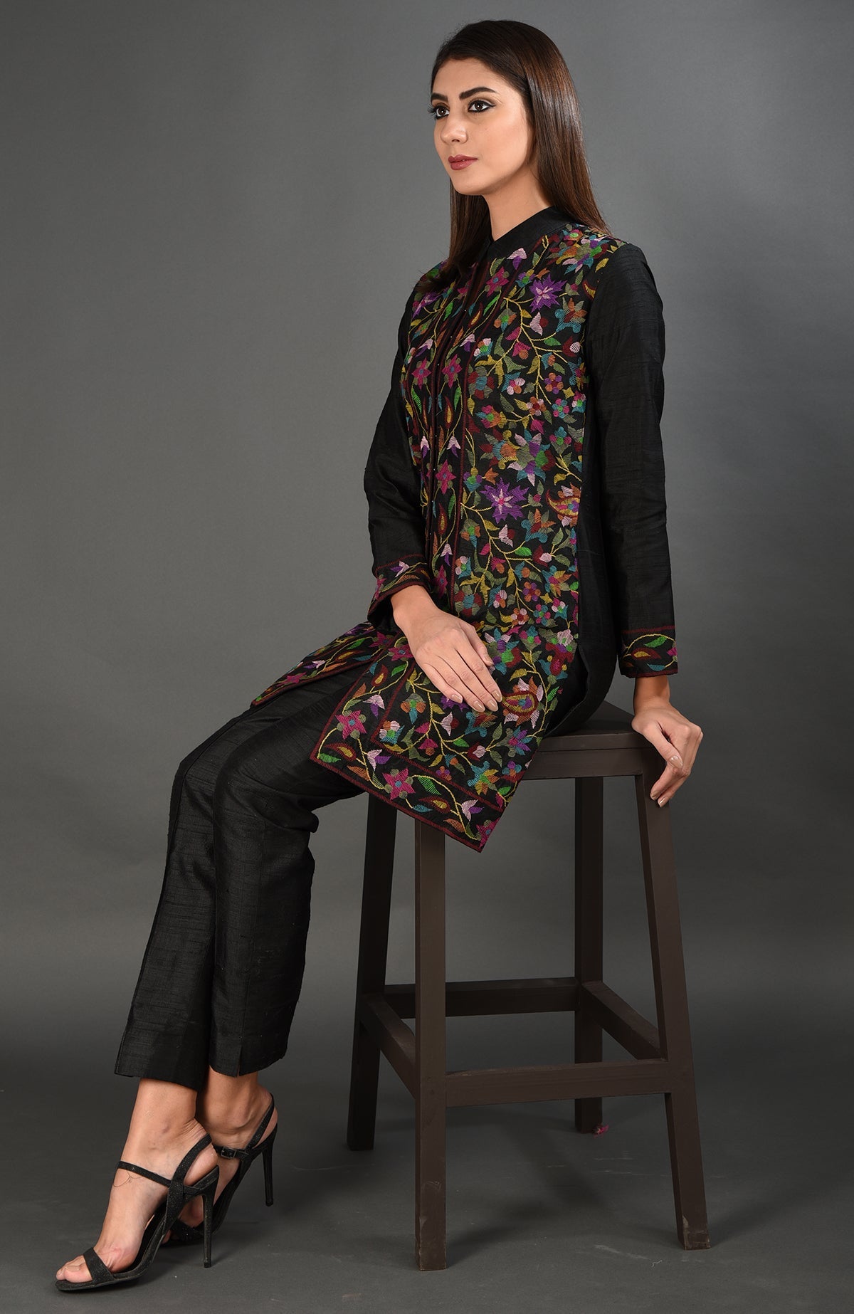 A woman sits wearing a black Kashmir Kani art embroidered jacket set.
