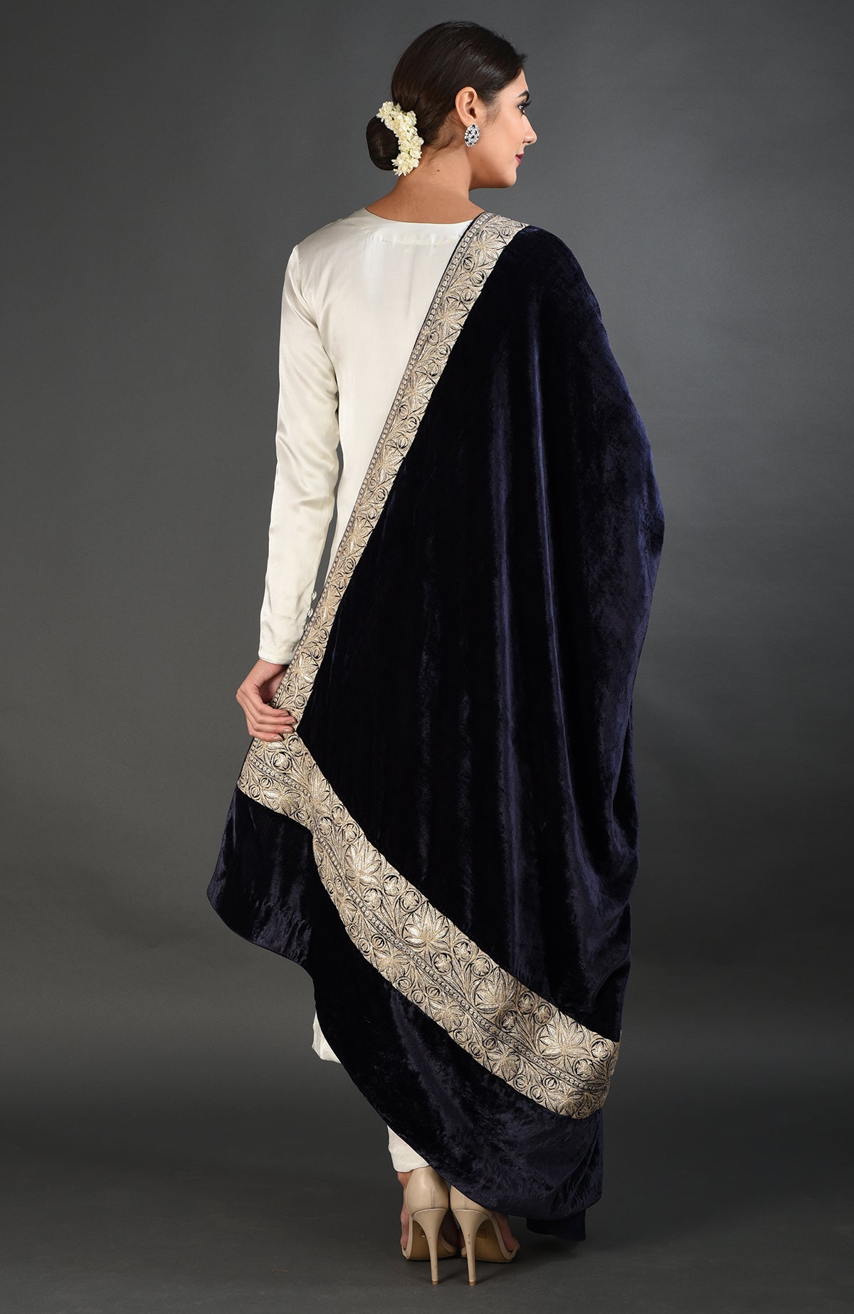 A woman in a white suit and nude heels holds a midnight blue velvet dupatta with silver embroidery.