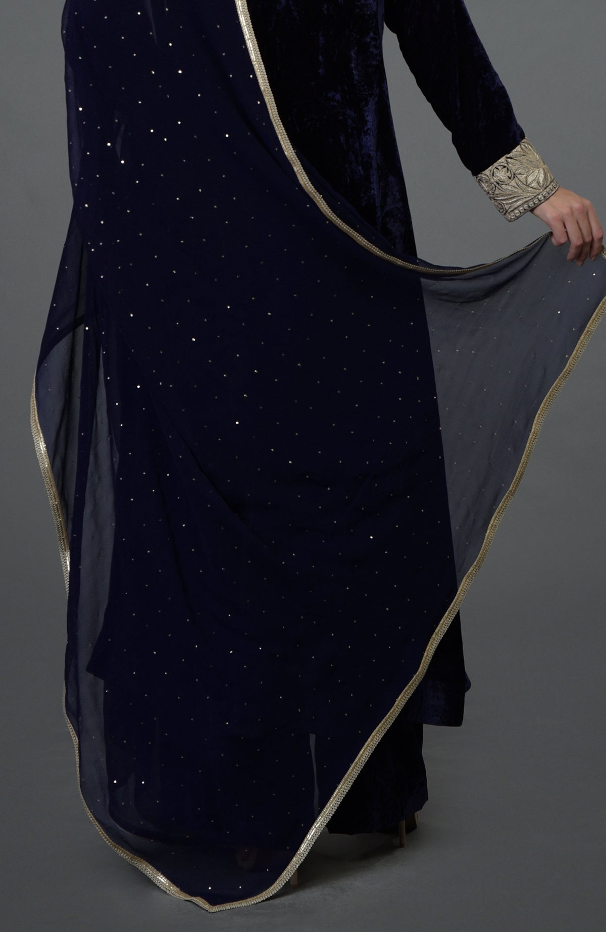 A person in a midnight blue velvet suit with an embroidered cuff and a sheer dupatta.