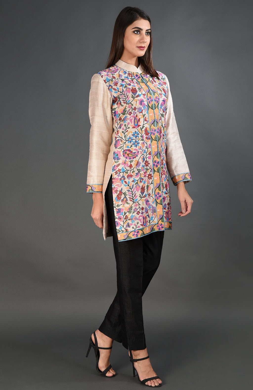 A woman models a creamy beige jacket with colorful floral embroidery and black pants.