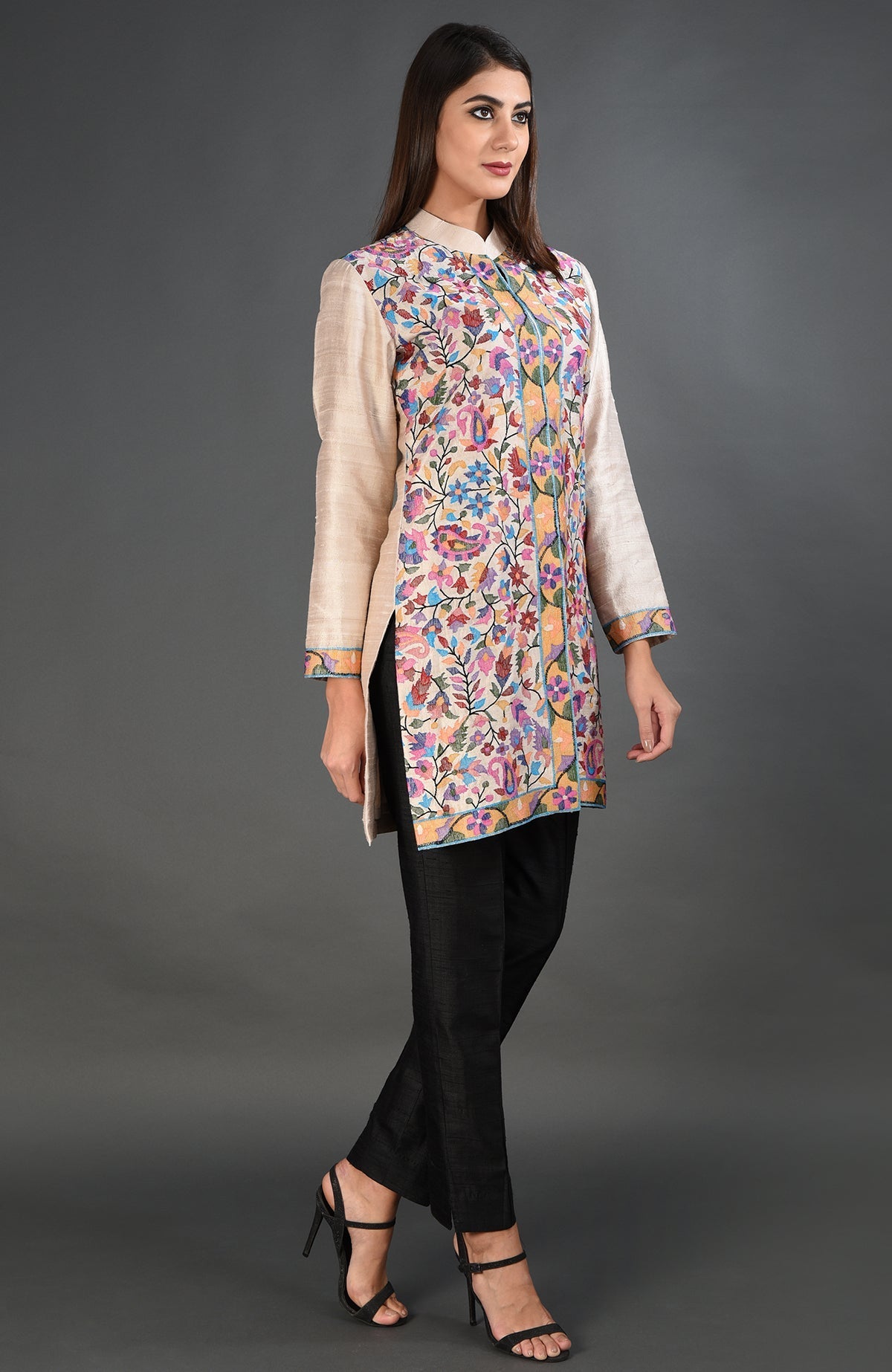 A woman models a creamy beige jacket with colorful floral embroidery and black pants.