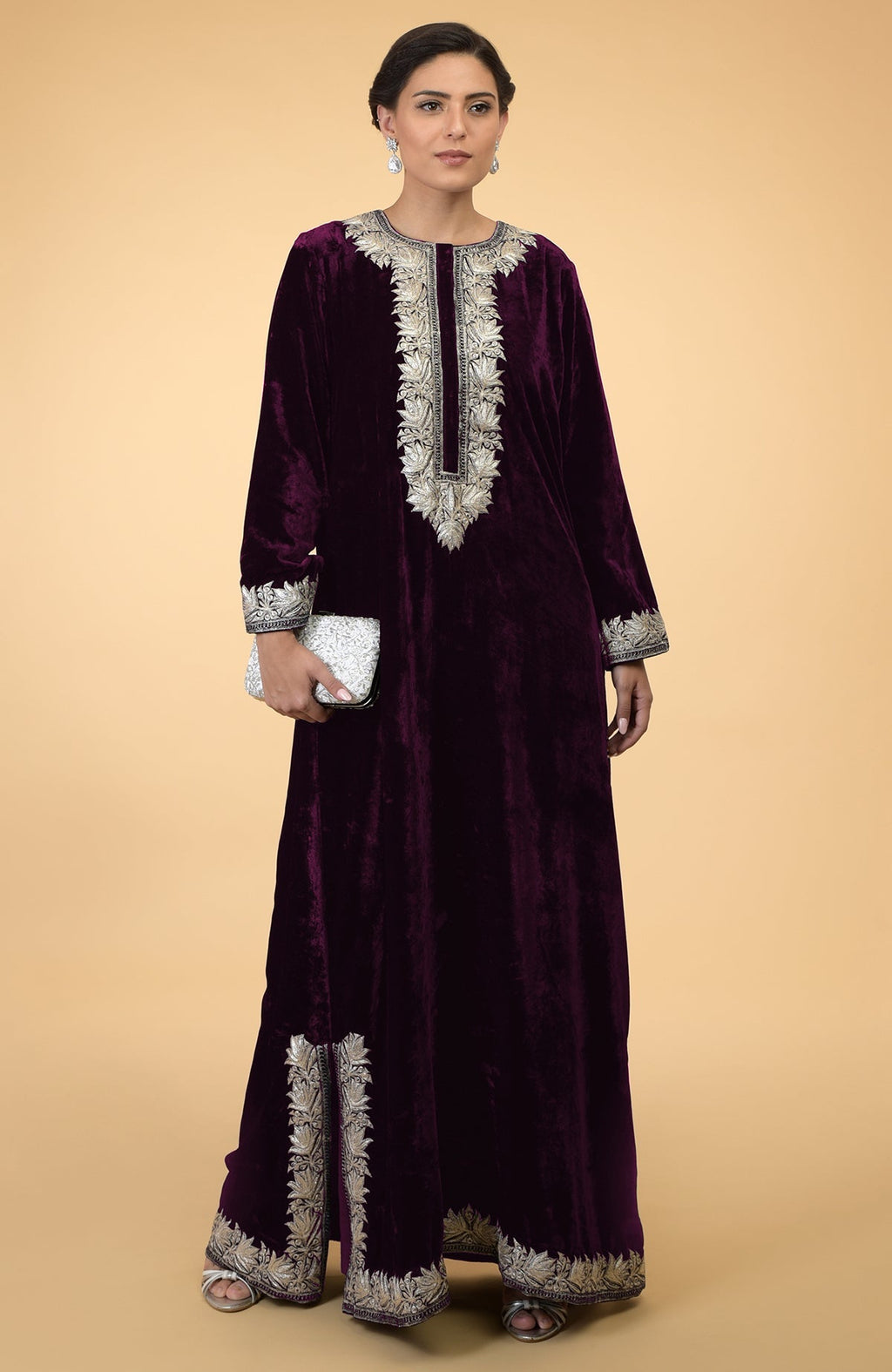A woman models a plum velvet kaftan with silver Kashmir embroidery and a white clutch.