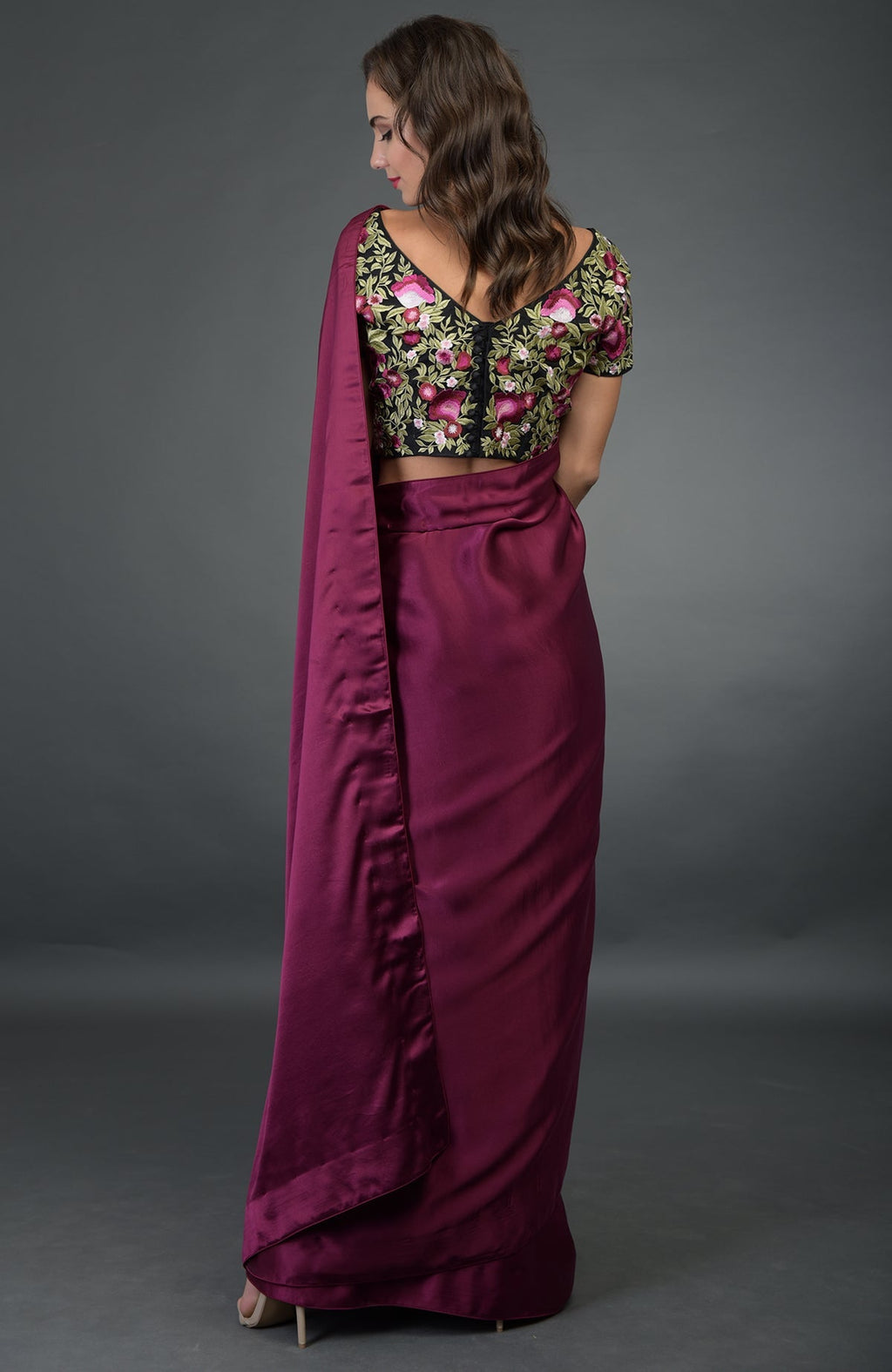 A woman models a plum saree with a black floral embroidered blouse from the back.