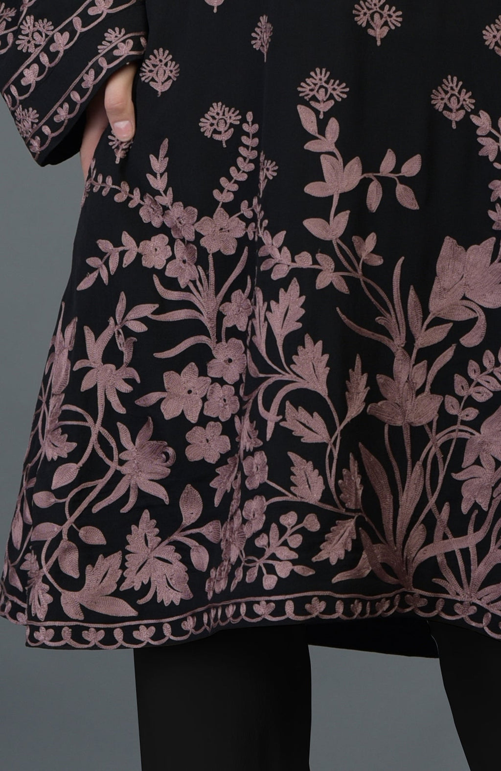 A black tunic with mauve Parsi floral embroidery and a hand in the pocket.