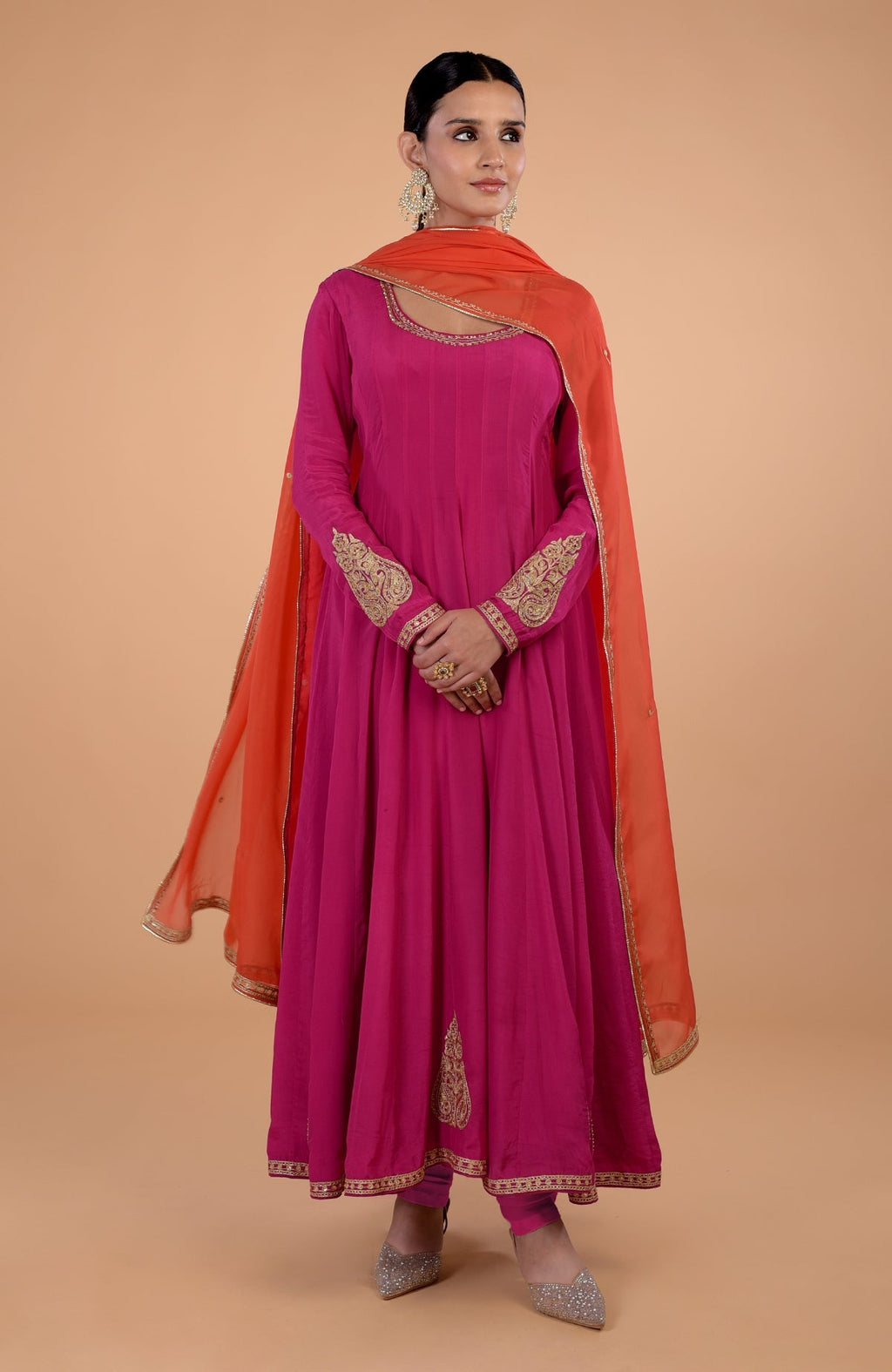 A woman models a raspberry pink Anarkali set with an orange dupatta and gold embroidery.