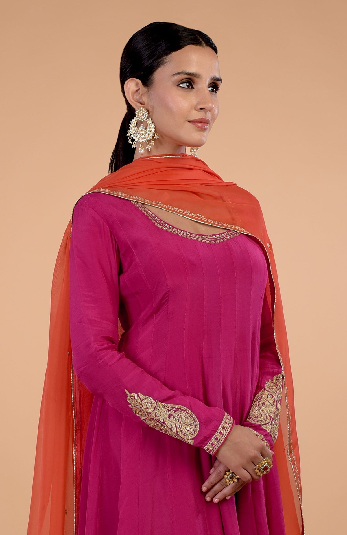 A woman in a raspberry pink Anarkali set with an orange dupatta and gold embroidery.