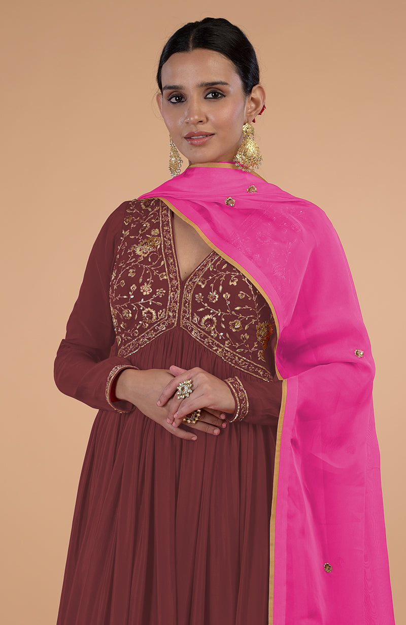 A woman models a marsala anarkali set with gold embroidery and a pink dupatta.