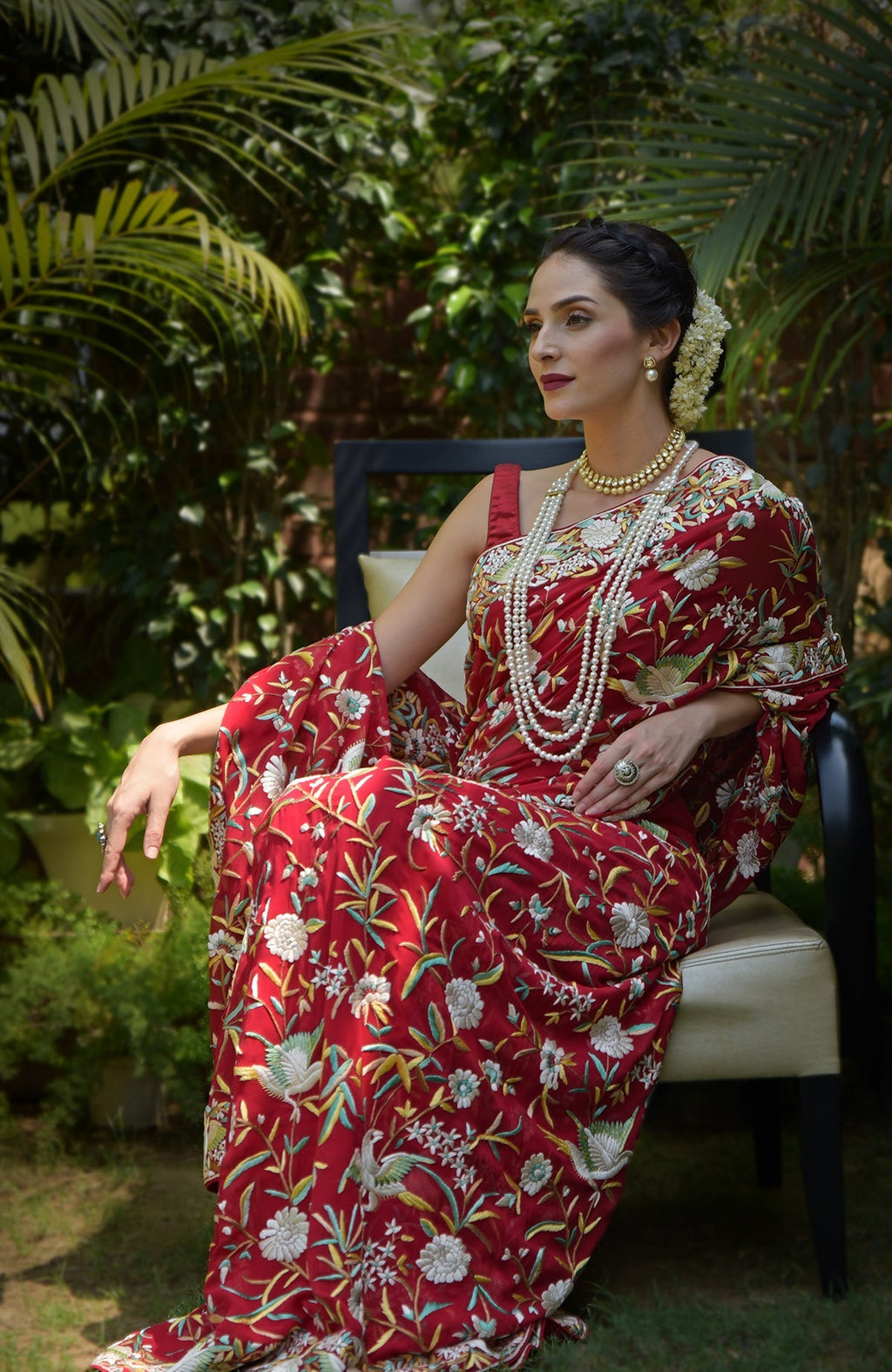 A woman sits wearing a red Parsi Gara hand-embroidered saree with floral and bird motifs.