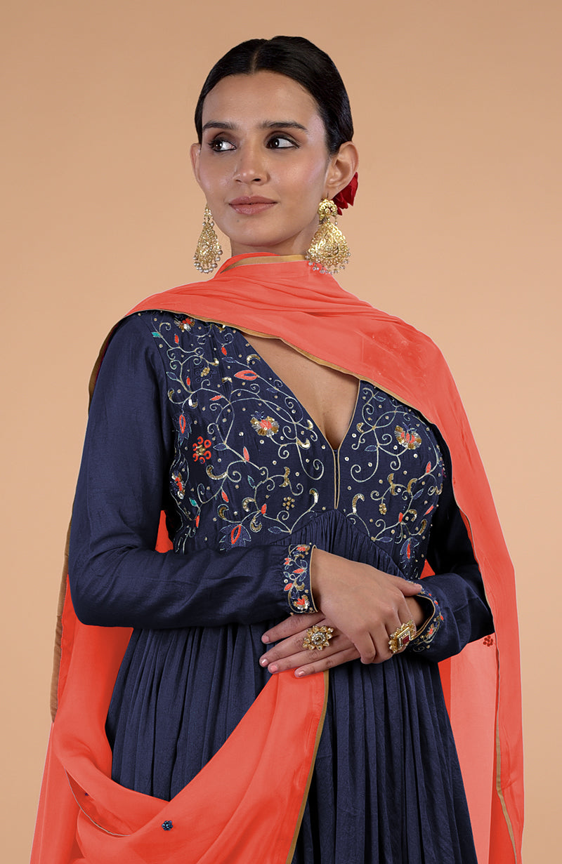 A woman in a navy blue anarkali with orange dupatta and gold earrings.