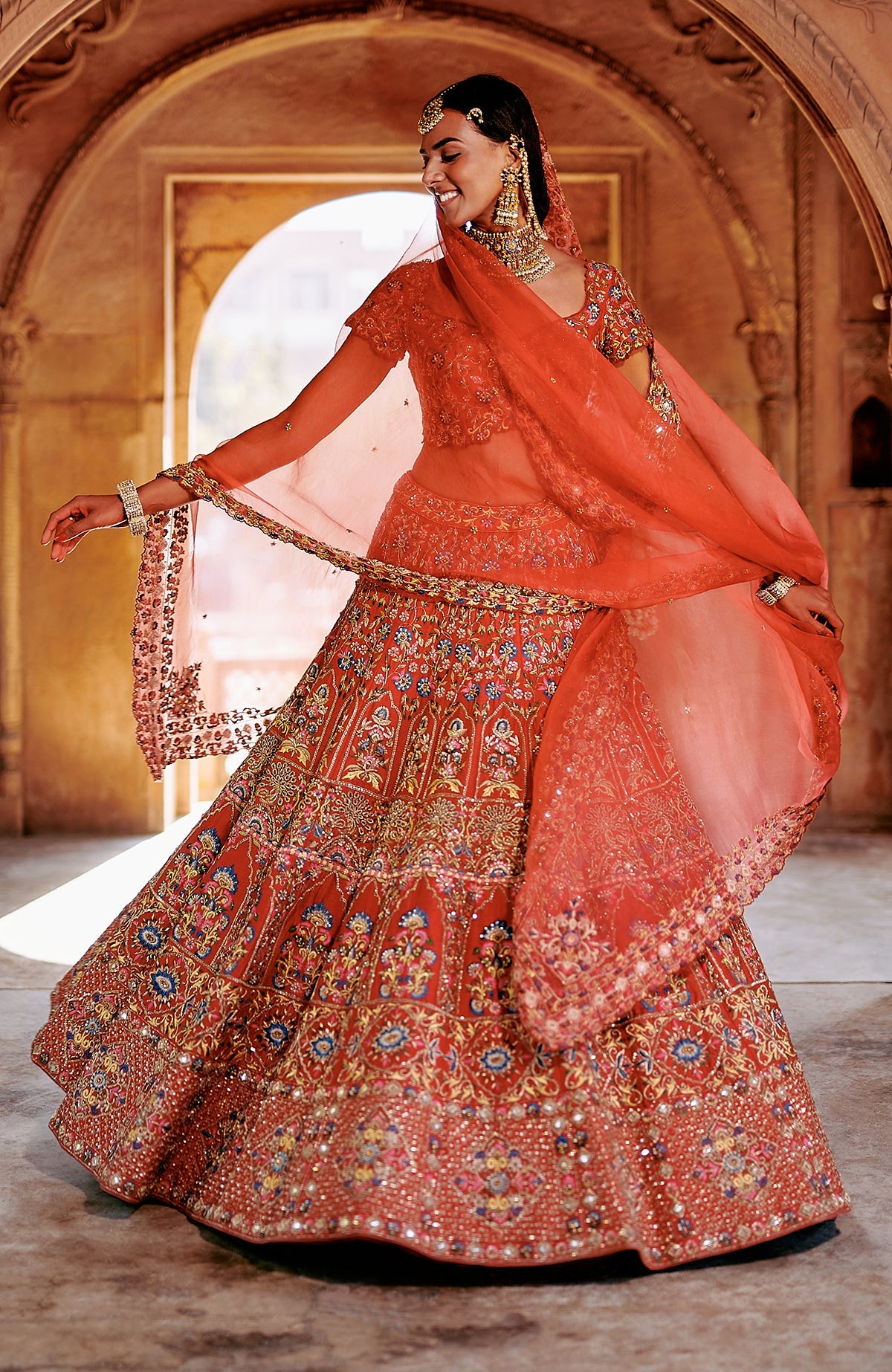 Red lehenga set with multicolor embroidery, gold zari, mirror work, and peacock feather tassels.