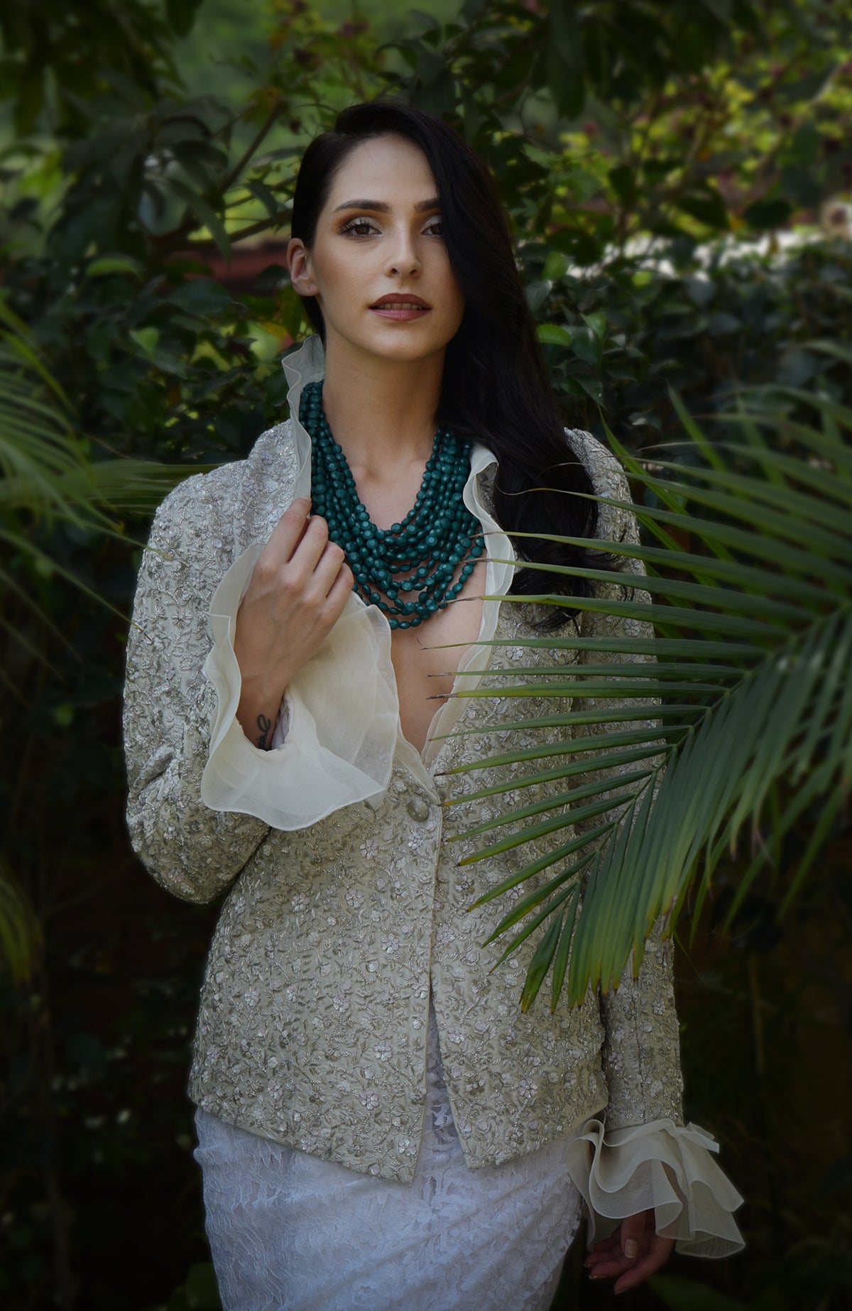 Woman wears a white sequin and bead embroidered jacket with a green beaded necklace.