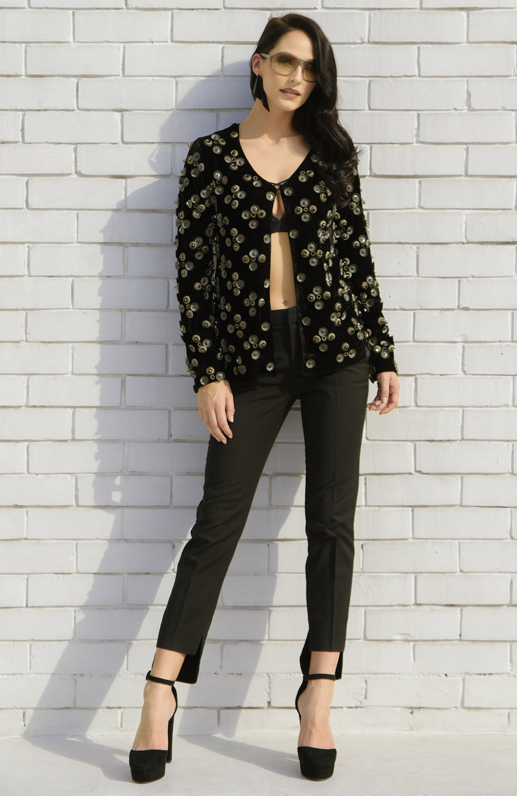 A woman models a black velvet jacket with metal studs, black trousers, and black heels.