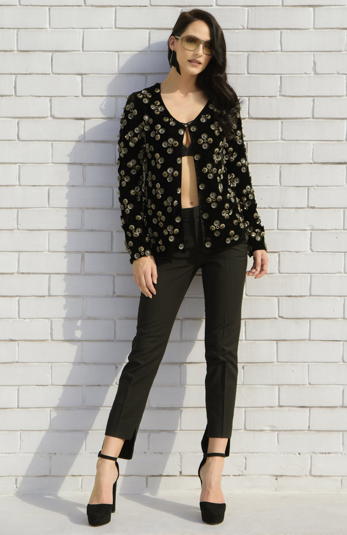 A woman models a black velvet jacket with metal studs, black trousers, and black heels.